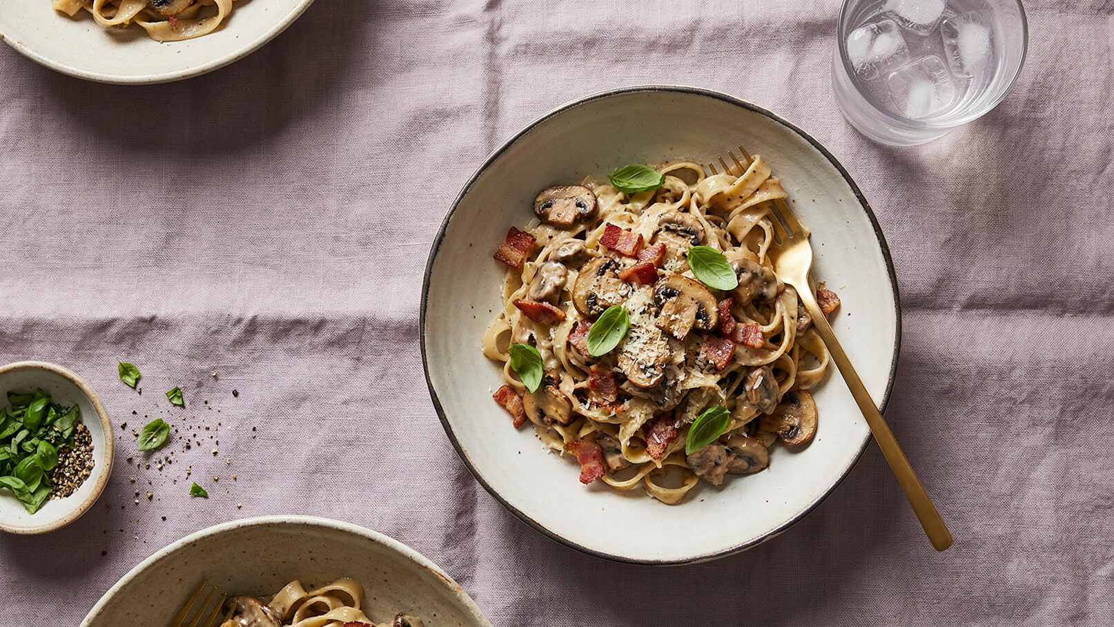 Ein Teller mit Tagliatelle in einer cremigen Sauce, garniert mit Champignons, Speck und frischem Basilikum. Der Teller steht auf einem lila Tischcloth, daneben eine Schale mit gehacktem Basilikum und eine Tasse Wasser mit Eiswürfeln.