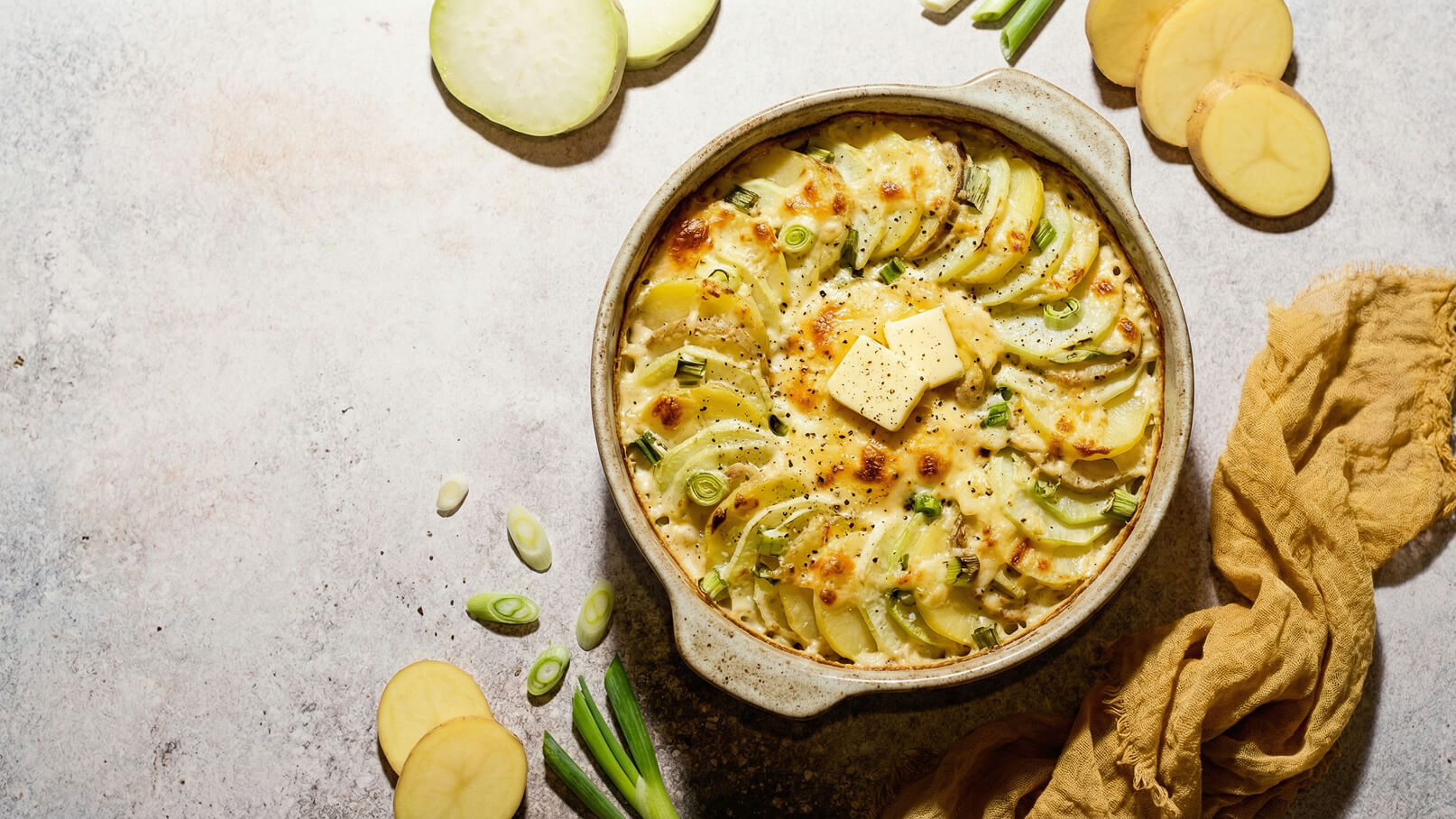 Ein runder Auflauf mit geschichteten Kartoffeln und Zucchini, überbacken mit Käse. Obenauf liegt ein Stück Butter. Neben dem Auflauf sind einige frische Frühlingszwiebeln und Kartoffelscheiben angerichtet. Der Hintergrund ist schlicht und setzt die Speise in den Fokus.
