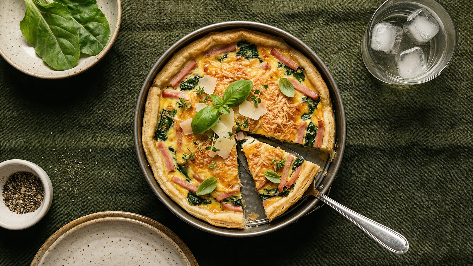 Eine frisch gebackene Quiche mit Spinat, Schinken und Käse. Ein Stück wurde herausgeschnitten und mit frischem Basilikum sowie Parmesan dekoriert. Im Hintergrund gibt es eine Schale mit frischem Spinat und ein Glas Wasser mit Eiswürfeln.