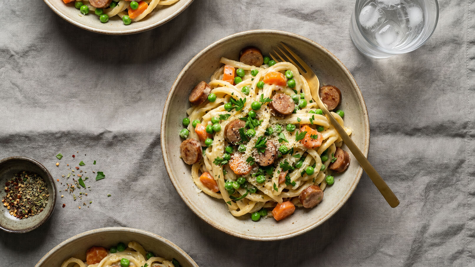 Ein Teller mit Tagliatelle, garniert mit Erbsen, Karotten und Scheiben von Wurst. Die Pasta ist in einer cremigen Sauce und mit frischer Petersilie bestreut. Im Hintergrund befinden sich weitere Teller und ein Glas Wasser mit Eiswürfeln.