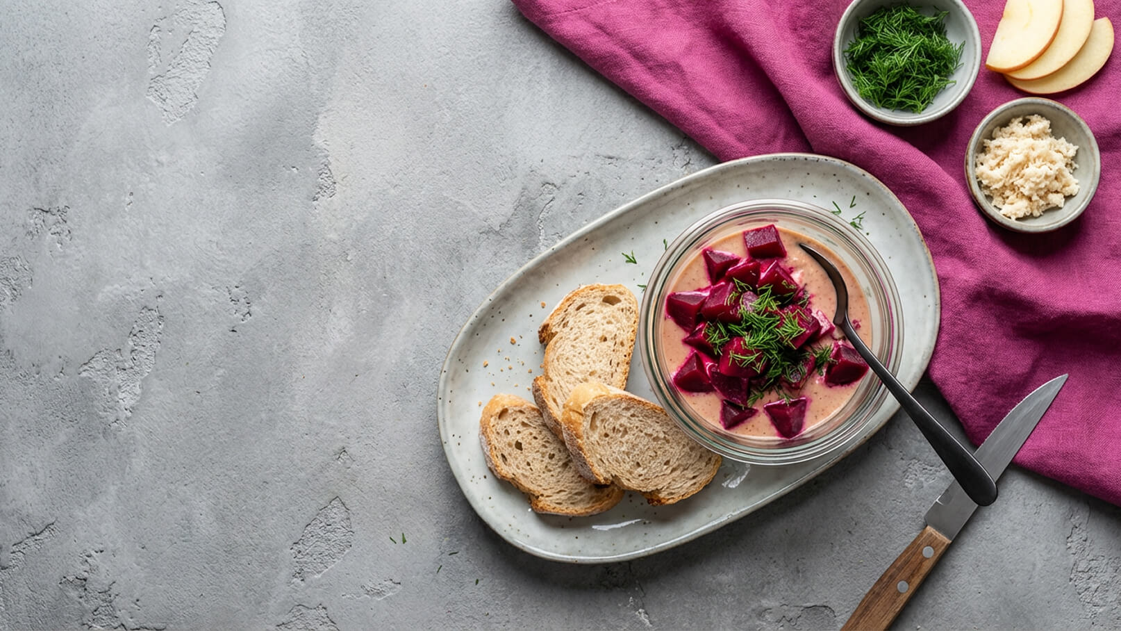 Ein Teller mit einer Portion Rote-Bete-Aufstrich, garniert mit frischen Kräutern, steht neben mehreren Scheiben Vollkornbrot. Im Hintergrund sind kleine Schalen mit weiteren Beilagen sowie ein Lappen in dunkler Farbe sichtbar. 
