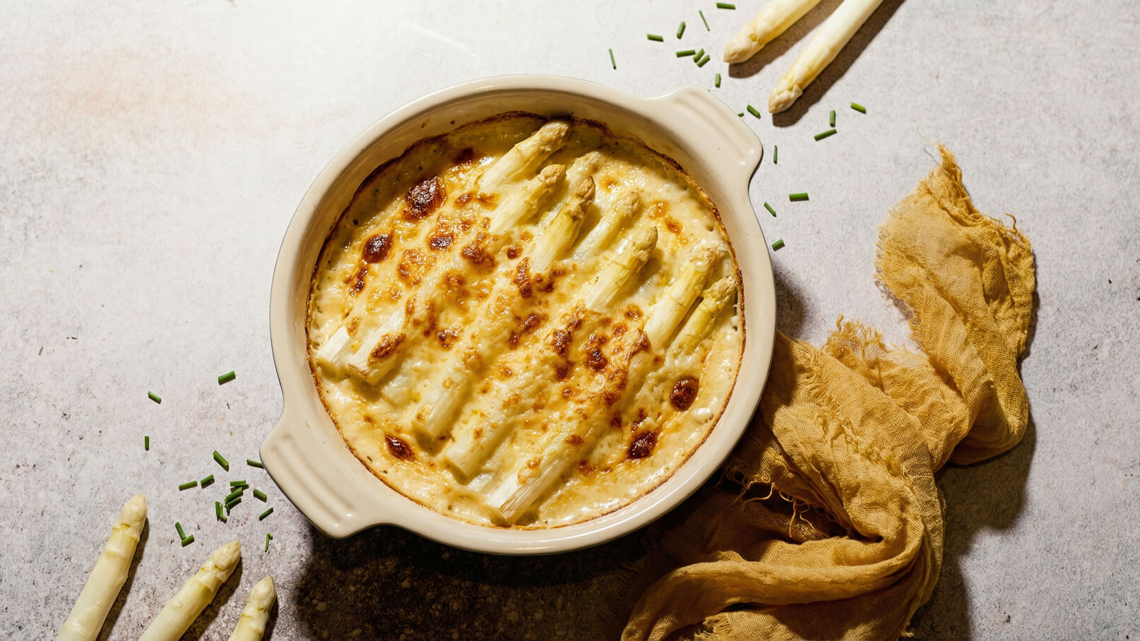 Ein Auflauf mit weißen Spargelstangen ist goldbraun überbacken und in einer hellen, runden Auflaufform. Um die Form herum liegen frische, rohe Spargelstangen und Schnittlauch. Der Hintergrund ist in neutralen Farbtönen gehalten.