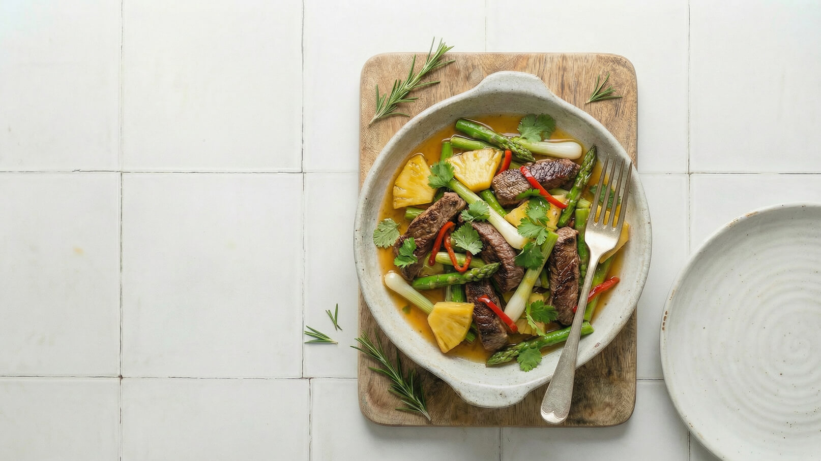 Eine Schüssel mit gebratenem Rindfleisch, grünem Spargel und Ananasstücken, garniert mit frischem Koriander und roten Chilischoten. Die Schüssel steht auf einem Holzbrett neben einem leeren Teller. Der Hintergrund ist schlicht und weiß gefliest.