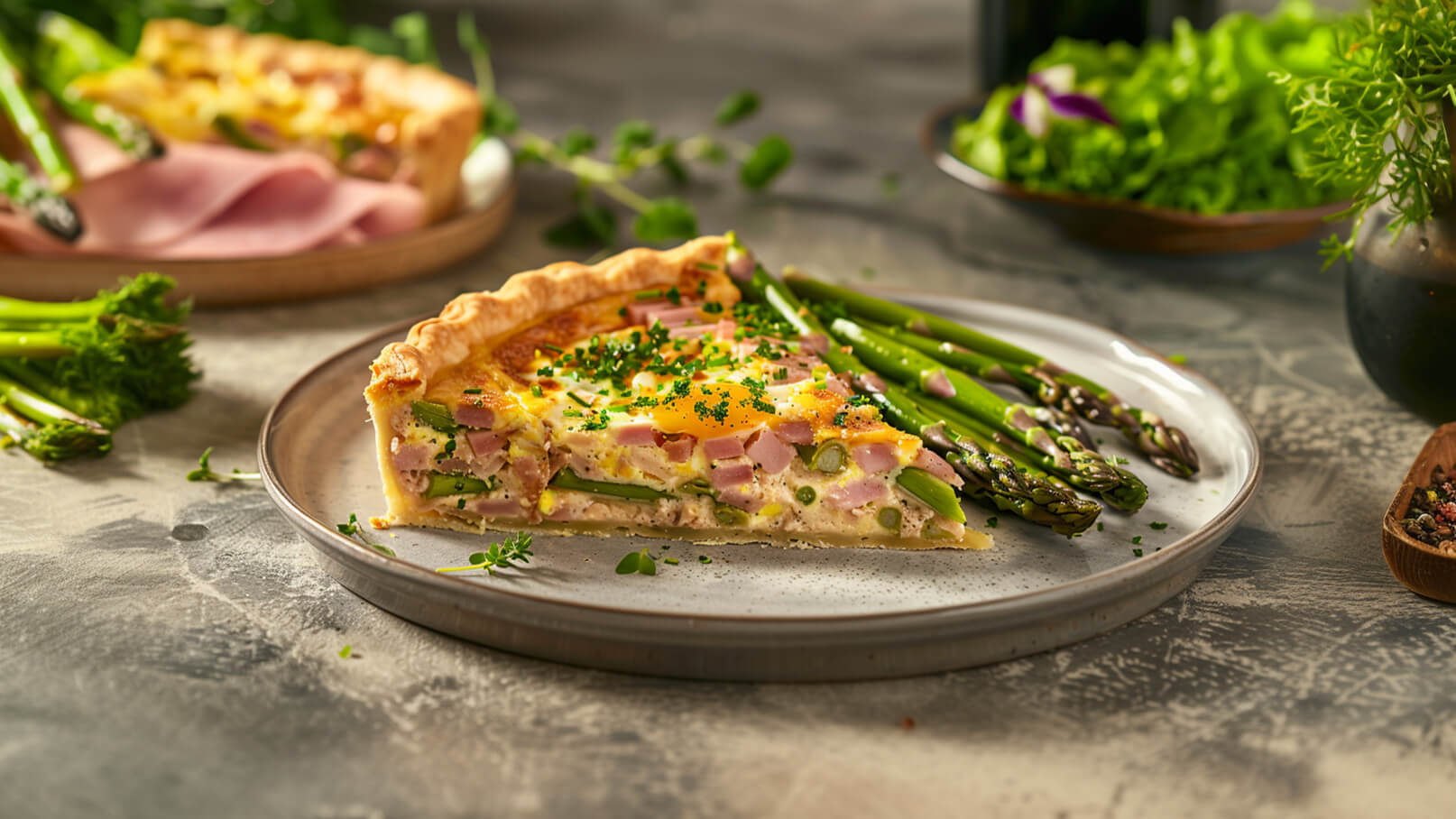 Ein Teller mit einer Quiche, gefüllt mit Schinken, grünem Spargel und einer Eierschicht. Die Quiche hat eine goldene, knusprige Kruste und wird mit frischem Schnittlauch garniert. Neben dem Teller liegen gekochte grüne Spargelstangen.
