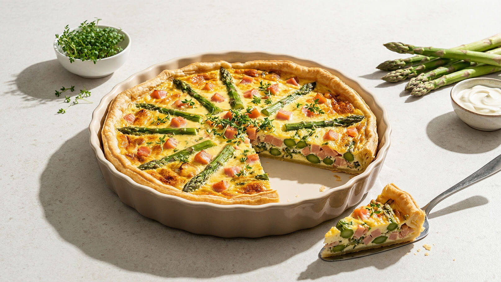Ein gefüllter Quiche ist auf einem hellen Tisch platziert. Die Quiche enthält grüne Spargelstücke, Schinken und frische Kräuter. Ein Stück wurde herausgenommen und liegt auf einem Teller. Neben der Quiche befindet sich eine kleine Schüssel mit saurer Sahne und frische Spargelstangen.