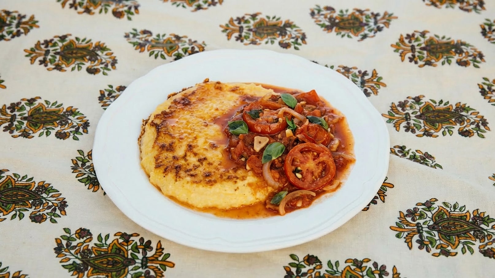 Ein Teller mit cremigem Polenta, der leicht gebräunt ist, wird mit einer aromatischen Tomatensauce garniert. Die Sauce enthält frische Tomaten, Zwiebeln und Basilikum. Der Hintergrund zeigt ein gemustertes Tischdeckenmuster.