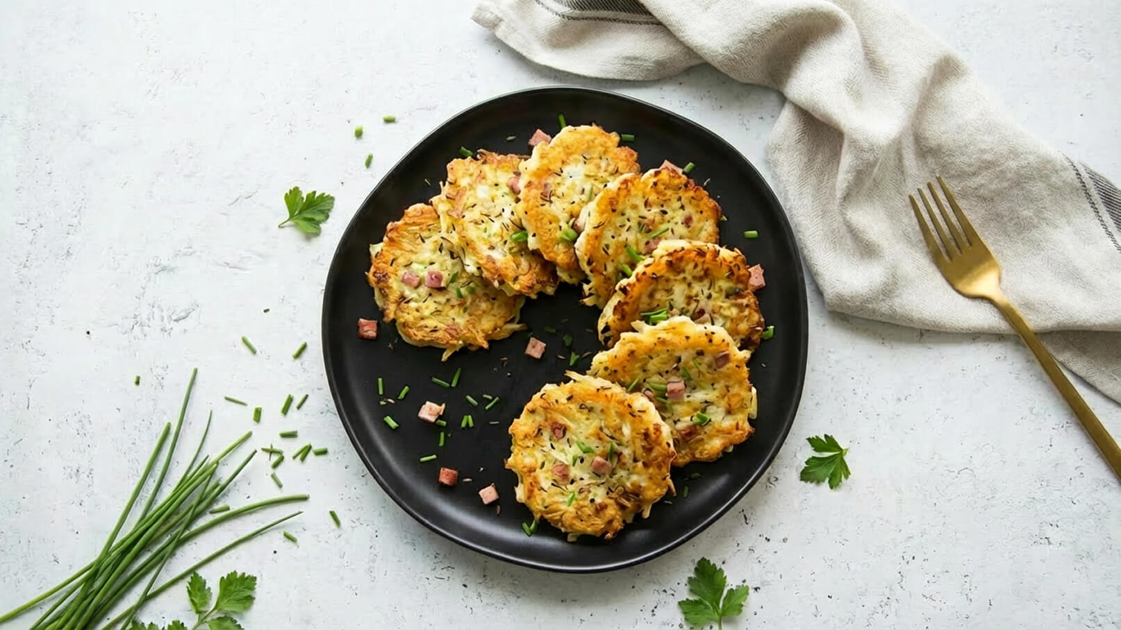 Ein schwarzes Teller mit mehreren goldbraunen Kartoffelpfannkuchen, die mit Schnittlauch und kleinen Schinkenstücken garniert sind. Einige frische Schnittlauchhalme und Kräuter sind um den Teller verteilt, im Hintergrund liegt ein helles Geschirrtuch und eine Gabel.