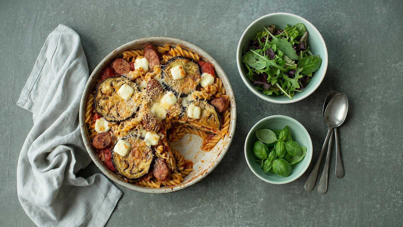Ein Teller mit Gemüse-Nudelauflauf, überbacken mit Käse, ist im Vordergrund zu sehen. Daneben befinden sich zwei Schalen: eine mit gemischtem grünen Salat und eine mit frischen Basilikumblättern. Ein graues Küchentuch liegt am Rand.