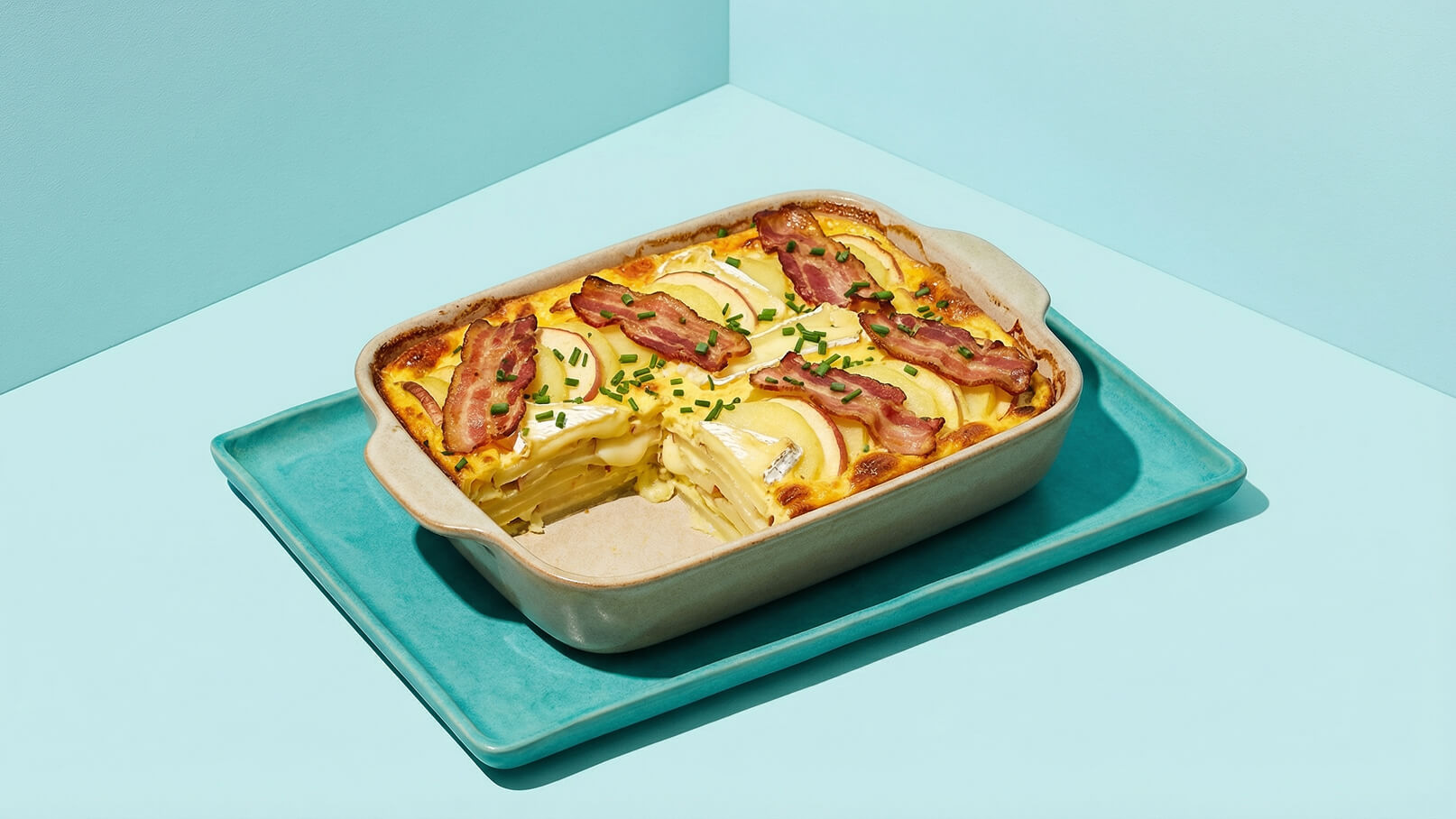 Auf einem hellblauen Teller liegt eine Keramikform mit herzhaftem Auflauf. Der Auflauf ist geschichtet mit Kartoffeln, Käse und einer Schicht gebratenem Speck, garniert mit Schnittlauch. Ein Stück des Auflaufs wurde herausgenommen, sodass die Füllung sichtbar ist.