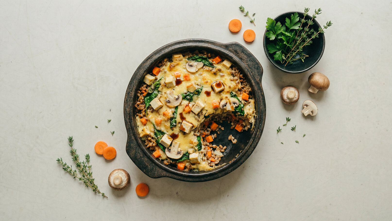Ein rustikales Gericht in einer schwarzen Auflaufform, das mit Spinat, Karotten, Pilzen und Käse belegt ist. Um die Form liegen einige frische Kräuter, Karottenscheiben und ganze Pilze. Das Gericht wirkt herzhaft und appetitlich.