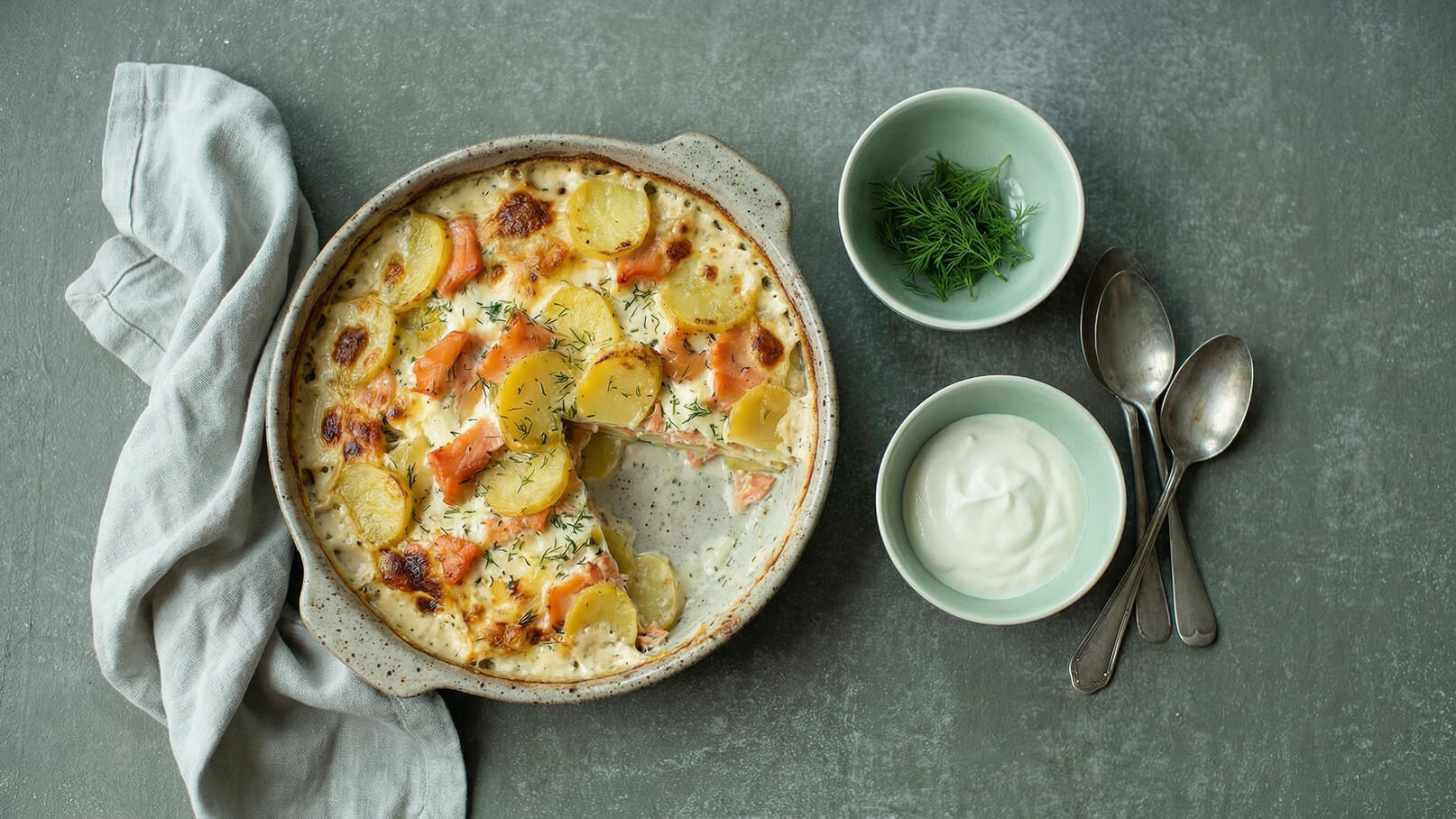 Ein Auflauf aus Kartoffeln und Lachs, mit einer goldbraunen Spitze, frisch gebacken in einer rundlichen Keramikform. Daneben befinden sich zwei kleine grüne Schalen, eine mit frischem Dill und die andere mit Joghurt, sowie drei silberne Löffel.