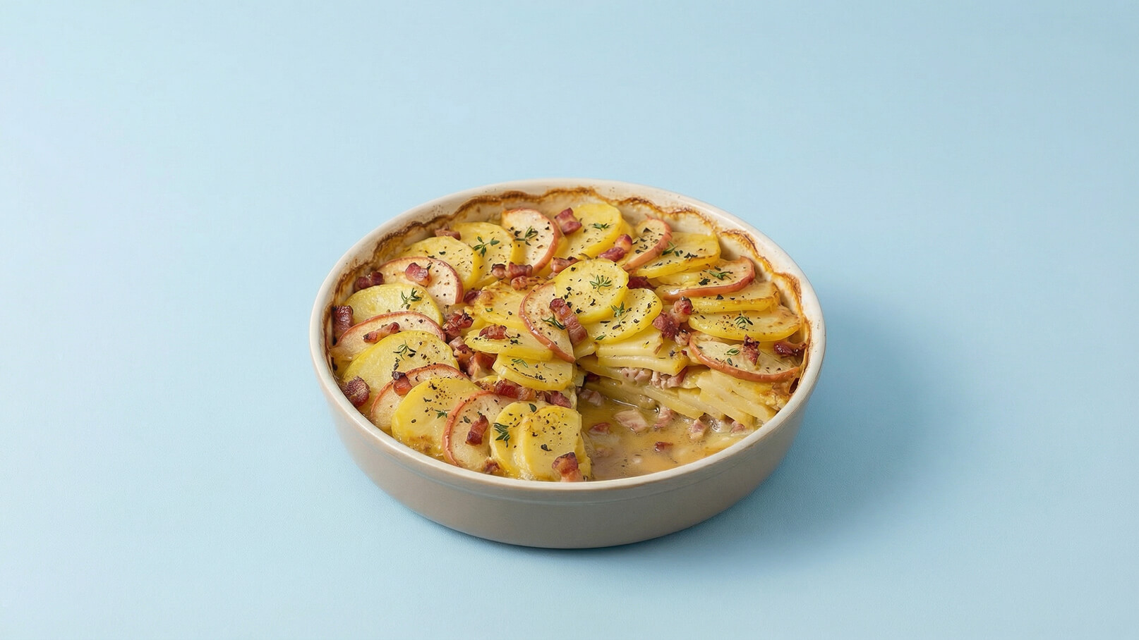 Eine runde Auflaufform mit geschichteten Kartoffeln, die mit Speck und Kräutern garniert sind. Die Kartoffeln sind goldbraun gebacken und haben eine appetitliche, knusprige Oberfläche. Der Hintergrund ist in einem sanften Blau gehalten.