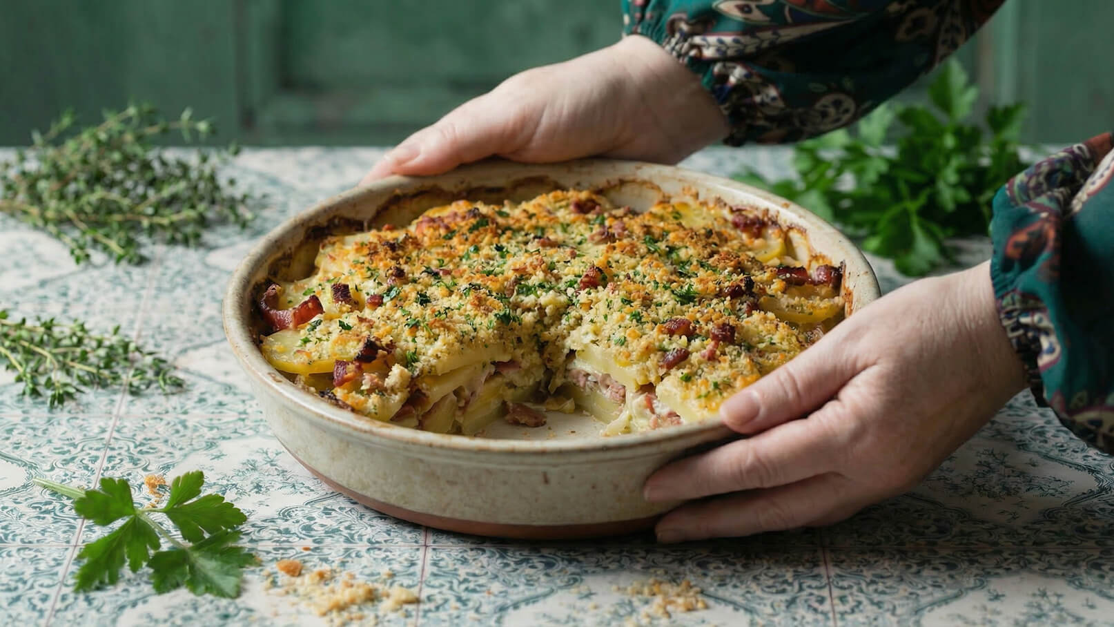 Eine Person hält eine flache Schüssel mit einem gratinierten Kartoffelgericht, das goldbraun und knusprig aussieht. Das Gericht enthält Schichten von Kartoffeln und möglicherweise Schinken, garniert mit Kräutern. Um die Schüssel herum sind frische Kräuter zu sehen.