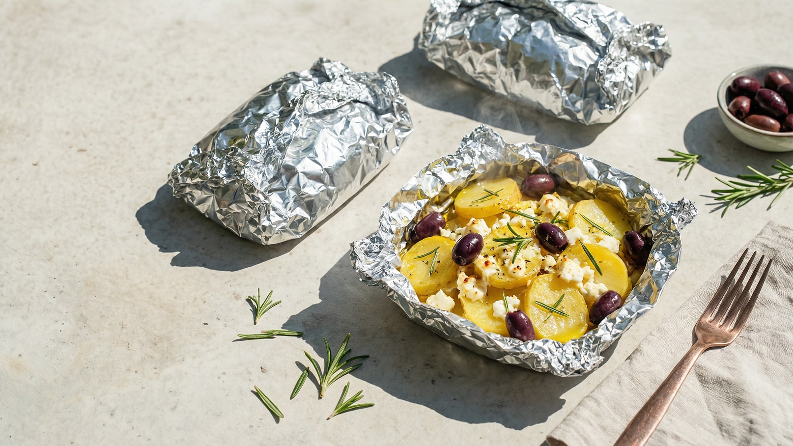 Ein silberner Alufolienbehälter enthält Kartoffeln, Feta, schwarze Oliven und frischen Rosmarin. Neben dem Behälter liegen zwei weitere eingewickelte Portionen in Alufolie. Im Hintergrund befindet sich eine Schale mit weiteren schwarzen Oliven und eine Gabel auf einem Serviette.