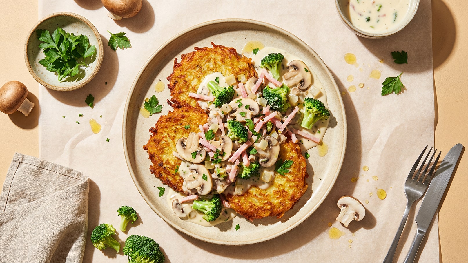 Ein Teller mit knusprigen Kartoffelpuffern, belegt mit einer cremigen Sauce, Champignons, Brokkoli und Schinken. Frische Petersilie dekoriert das Gericht. Neben dem Teller liegt eine Serviette und ein kleiner Schälchen mit einer weiteren Sauce.