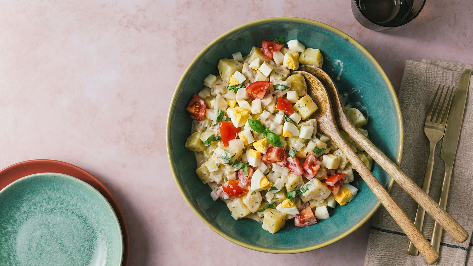 Eine große Schüssel mit einem bunten Salat, der Kartoffeln, Tomaten, Kräutern und kleinen Stückchen von hartgekochten Eiern enthält. Zwei Holzlöffel liegen in der Schüssel. Im Hintergrund sind zwei Teller und Besteck sichtbar.
