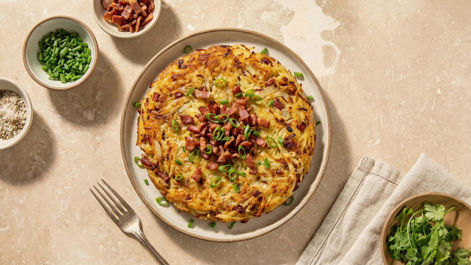 Ein runder Teller mit einer goldbraunen Kartoffelpfanne, belegt mit gewürfeltem Speck und frischen Frühlingszwiebeln. Daneben befinden sich kleine Schalen mit gehacktem Schnittlauch, Pfeffer und frischem Koriander. Eine Gabel liegt bereit, um das Gericht zu servieren.