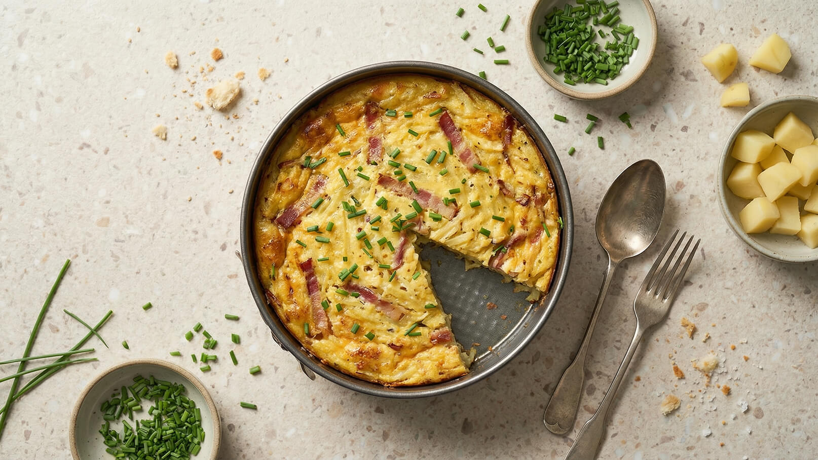 Ein runder Auflauf aus Kartoffeln, Eiern und Schinken, garniert mit frischem Schnittlauch. Ein Stück wurde entnommen, wodurch die Textur und die Zutaten sichtbar werden. Neben dem Auflauf liegen frische Schnittlauchröllchen und gewürfelte Kartoffeln auf einem hellen Tisch.