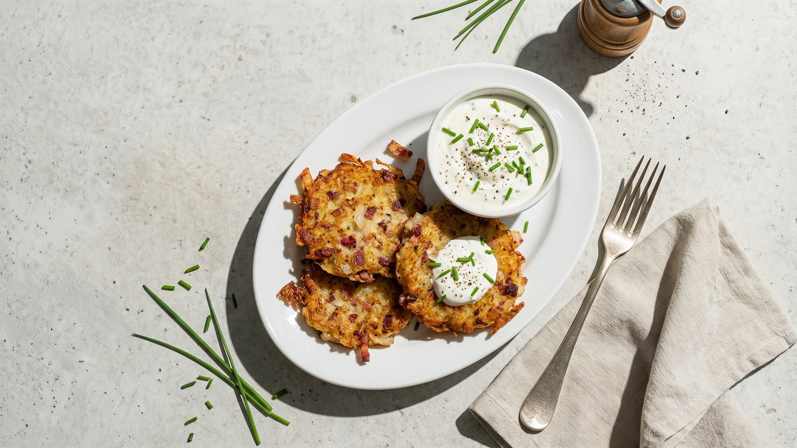 Ein Teller mit drei knusprigen Kartoffelpuffern, garniert mit einem Klecks saurer Sahne und Schnittlauch. Daneben steht eine Schüssel mit weiterer saurer Sahne, ebenfalls mit Schnittlauch bestreut. Eine Gabel und ein Tuch liegen neben dem Teller.