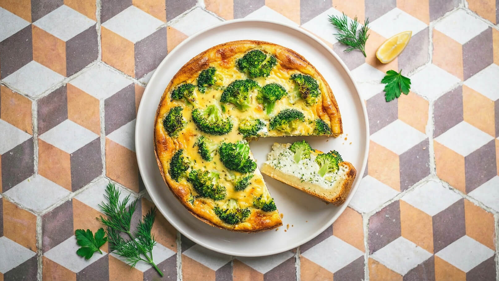 Ein rundes Quiche mit Brokkoli und einer goldbraunen Kruste liegt auf einem weißen Teller. Ein Stück ist herausgeschnitten, und frische Kräuter sowie eine Zitronenscheibe sind daneben platziert. Der Hintergrund zeigt ein geometrisches Muster in warmen Farben.