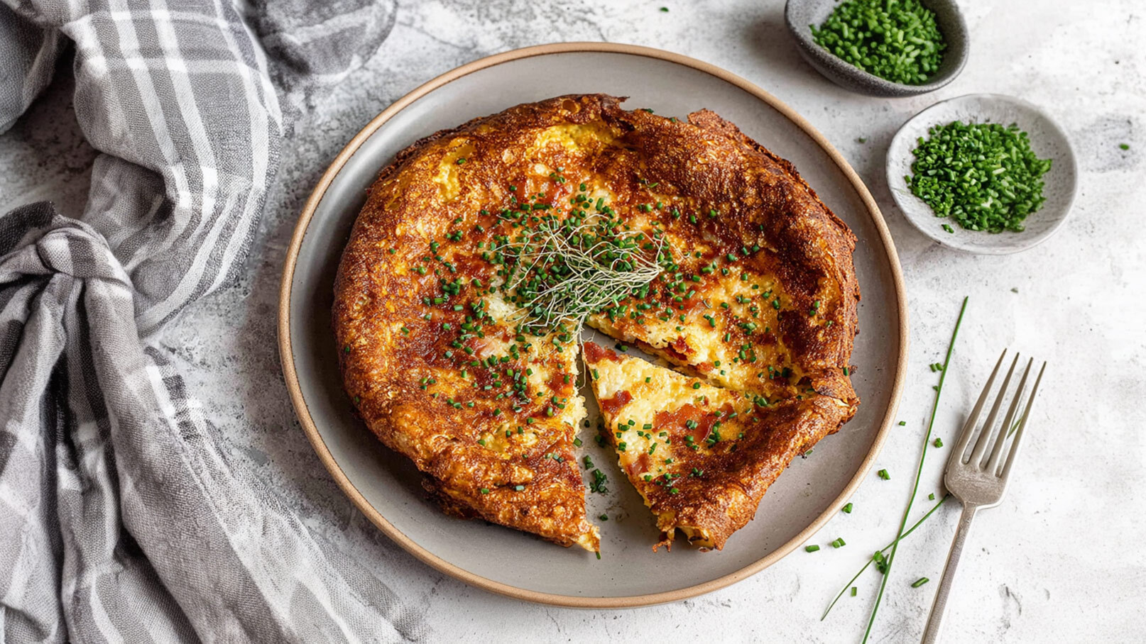 Ein Teller mit einer großen, goldbraunen Omelette, garniert mit frischem Schnittlauch. Ein Stück ist herausgeschnitten. Im Hintergrund befinden sich kleine Schalen mit extra Schnittlauch und eine Gabel. Ein kariertes Tuch liegt daneben.