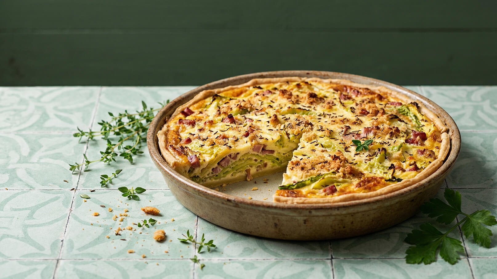 Ein runder, goldbrauner Auflauf mit einer knusprigen Kruste, gefüllt mit einer Mischung aus frischem Gemüse und Schinken. Ein Stück wurde herausgeschnitten, und einige Kräuter sind um den Auflauf herum verteilt. Der Hintergrund ist in sanften Grüntönen gehalten.