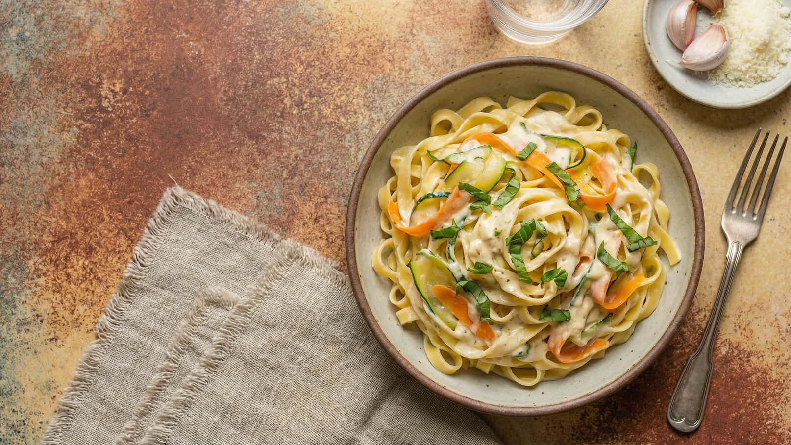 Ein Teller mit Pasta in einer cremigen Sauce, garniert mit feinen Streifen von Zucchini, Karotten und frischem Basilikum. Neben dem Teller liegt eine Gabel, und im Hintergrund sind eine kleine Glasschale mit Zutaten zu sehen.