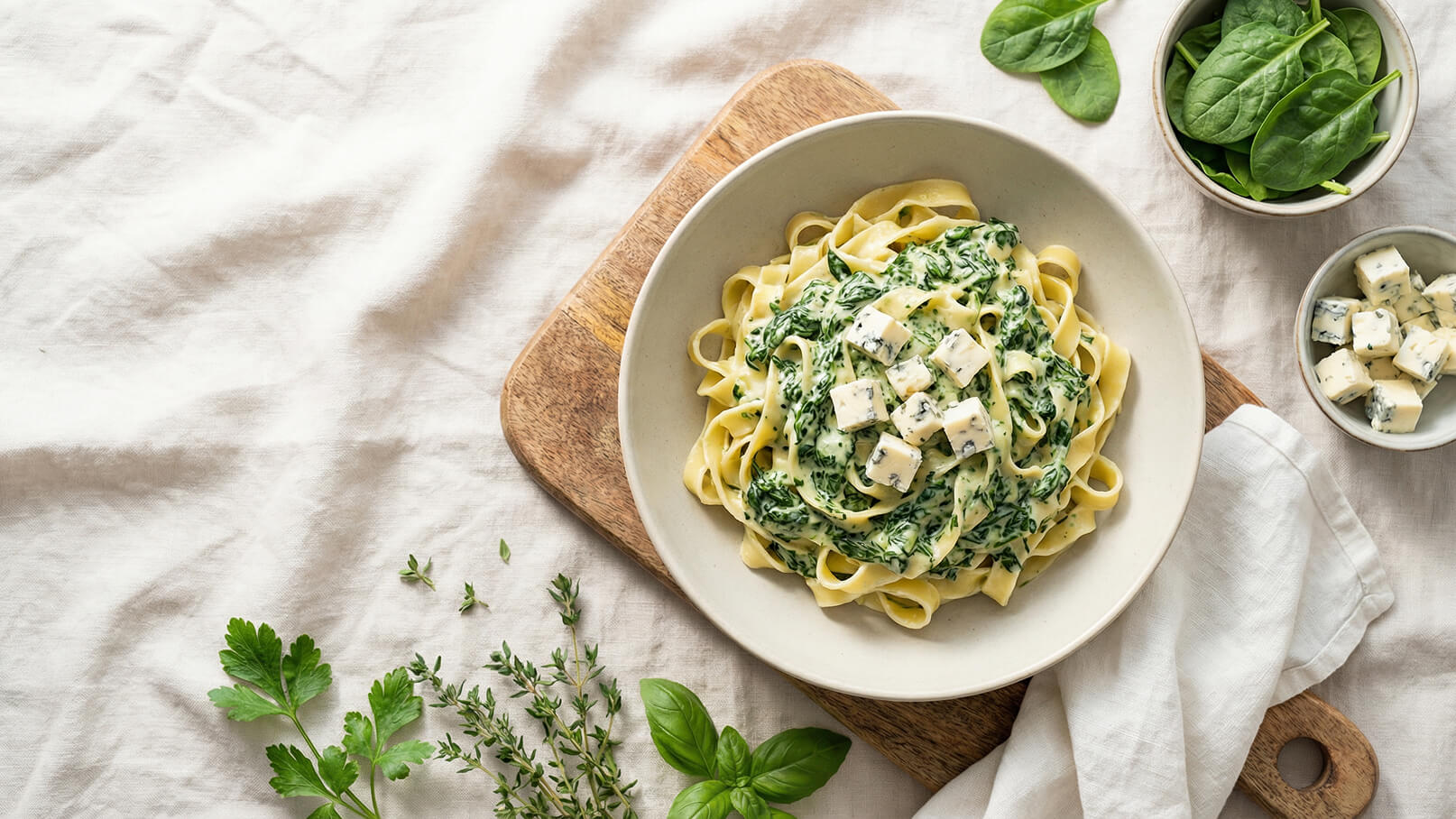 Ein Teller mit Bandnudeln in einer cremigen Spinatsoße, garniert mit würfelförmigem Käse, steht auf einem Holzbrett. Neben dem Teller liegen frische Spinatblätter und einige Kräuter. Der Hintergrund ist ein heller, strukturierter Stoff.