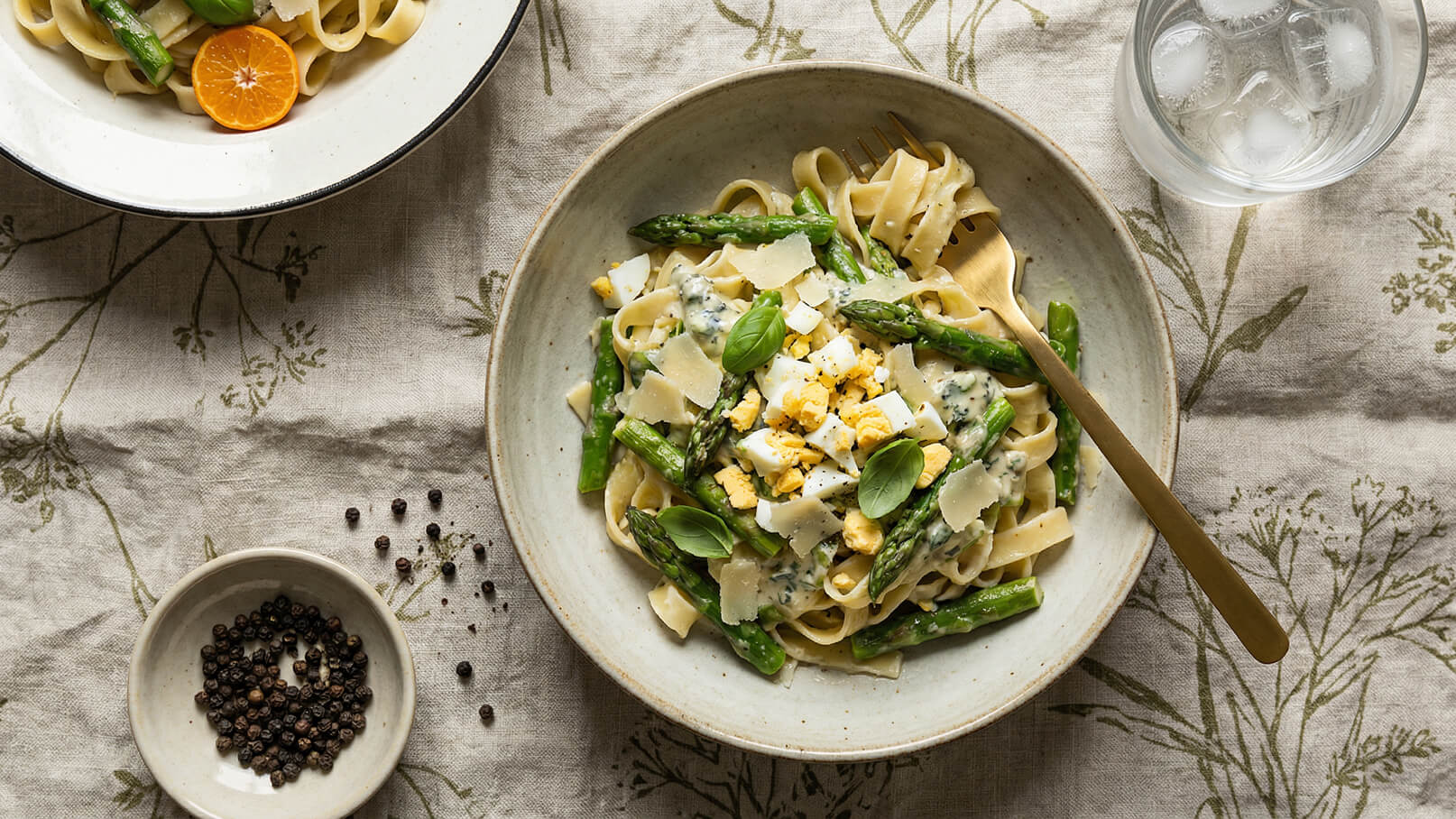 Eine Schüssel mit frischen Bandnudeln, garniert mit grünen Spargelstücken, gehacktem hartgekochten Ei, Parmesan und frischem Basilikum. Neben der Schüssel liegt eine Gabel. Im Hintergrund ist ein Glas mit Eiswasser und eine weitere Schüssel mit einer kleinen Portion Pfefferkörnern zu sehen.