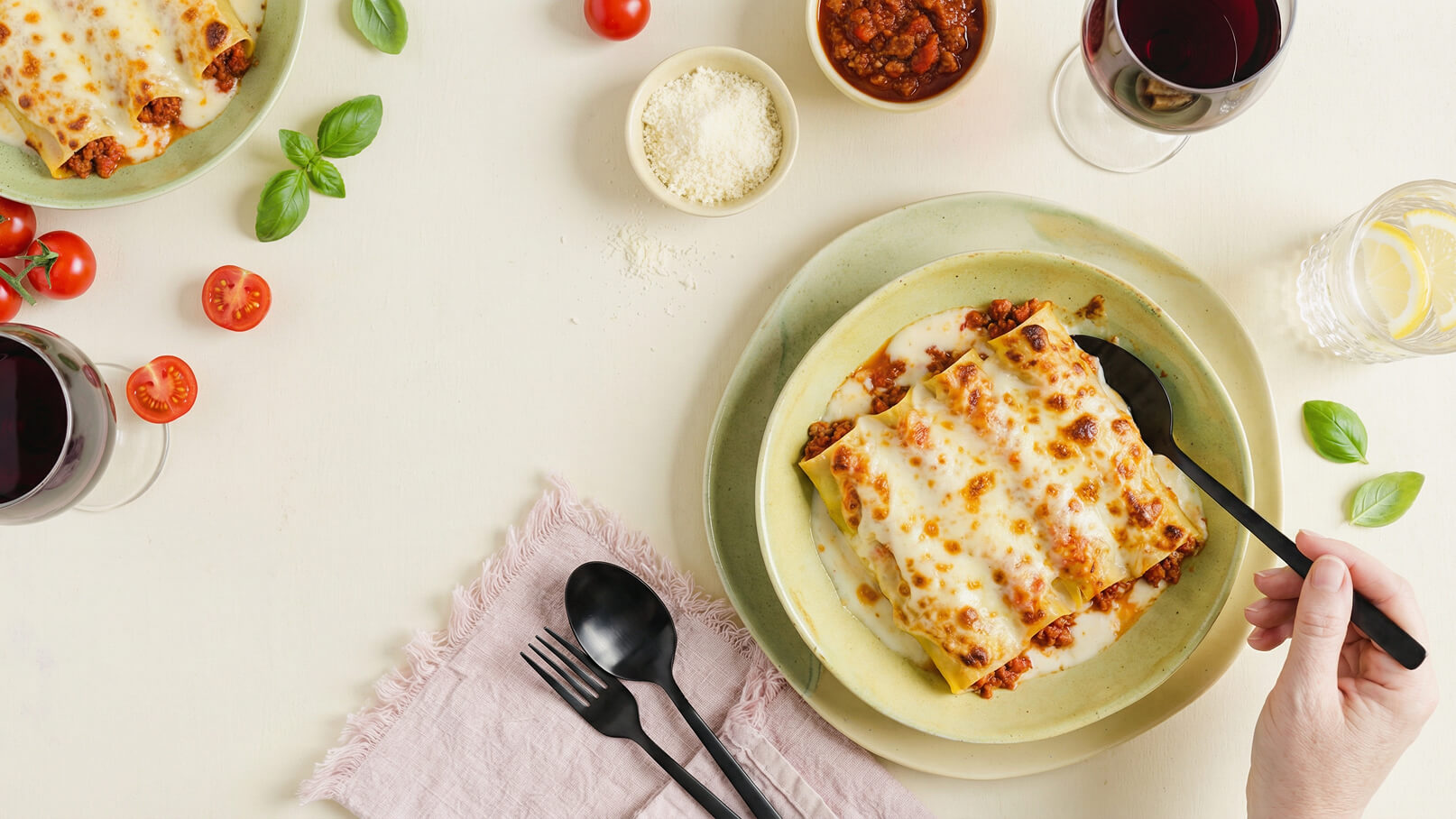 Ein Teller mit Lasagne, reichlich überbacken mit Käse, steht auf einem Tisch. Daneben sind KirschTomaten, ein Glas Rotwein und eine Schale mit Sauce. Eine Hand hält einen Löffel, bereit, die Lasagne zu servieren.