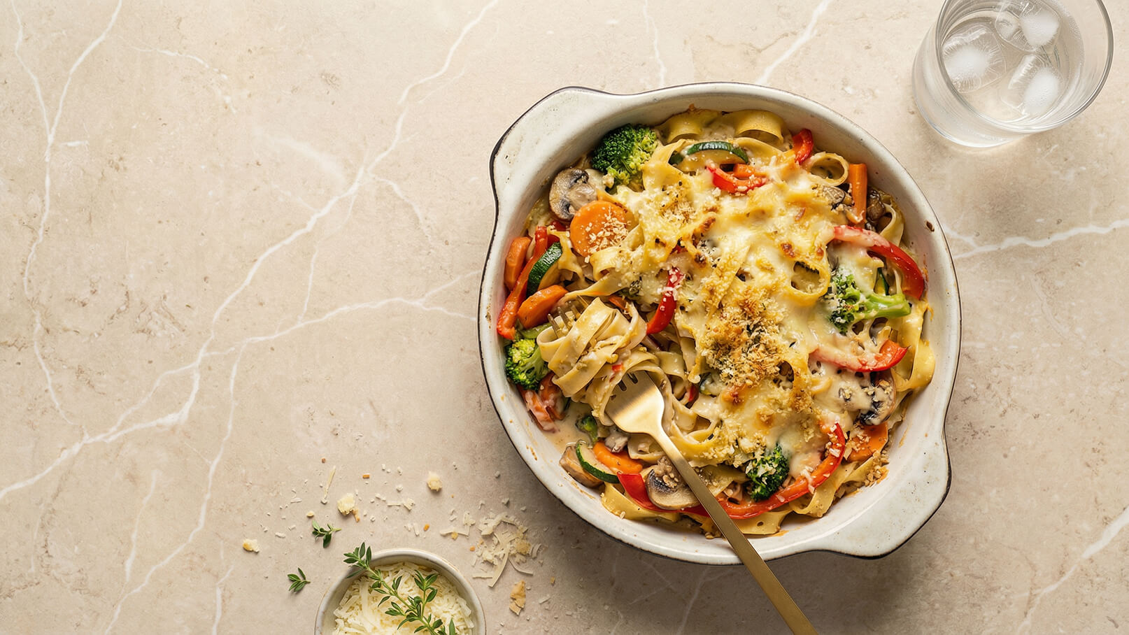 Ein reichhaltiges Nudelgericht in einer weißen Keramikschüssel. Es besteht aus Bandnudeln, buntem Gemüse wie Brokkoli, Paprika und Karotten, alles überbacken mit geschmolzenem Käse und einer knusprigen Panade. Ein Esslöffel steht zur Seite, während ein Glas Wasser daneben platziert ist.
