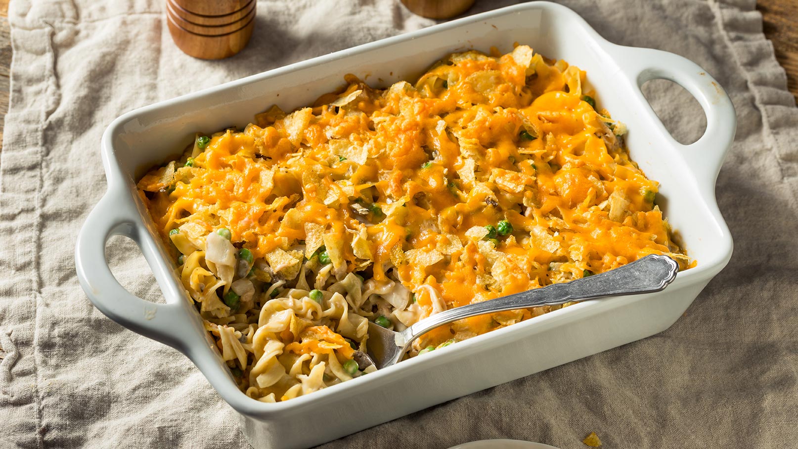 Stell dir eine weiße Auflaufform vor, prall gefüllt mit einem bunten Gemüse-Nudel-Auflauf. Die goldbraunen Nudeln sind von einer cremigen Sauce umhüllt und von frischem Gemüse wie Brokkoli, Paprika und Tomaten durchzogen. In der Form steckt ein silberner Löffel, bereit zum Servieren. Die Auflaufform steht auf einem weichen, beigefarbenen Tuch, was dem Bild eine einladende Atmosphäre verleiht.