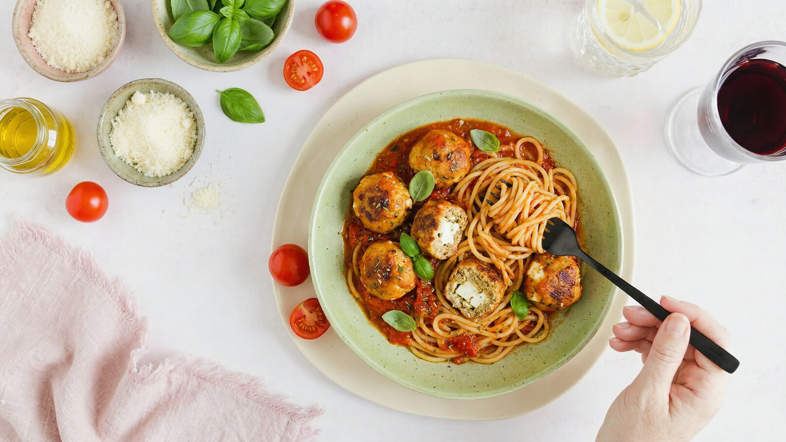 Ein Teller mit Spaghetti in Tomatensoße und Frikadellen, garniert mit frischem Basilikum. Eine Hand hält eine schwarze Gabel, um die Spaghetti zu probieren. Rote Kirschtomaten und Zutaten im Hintergrund.