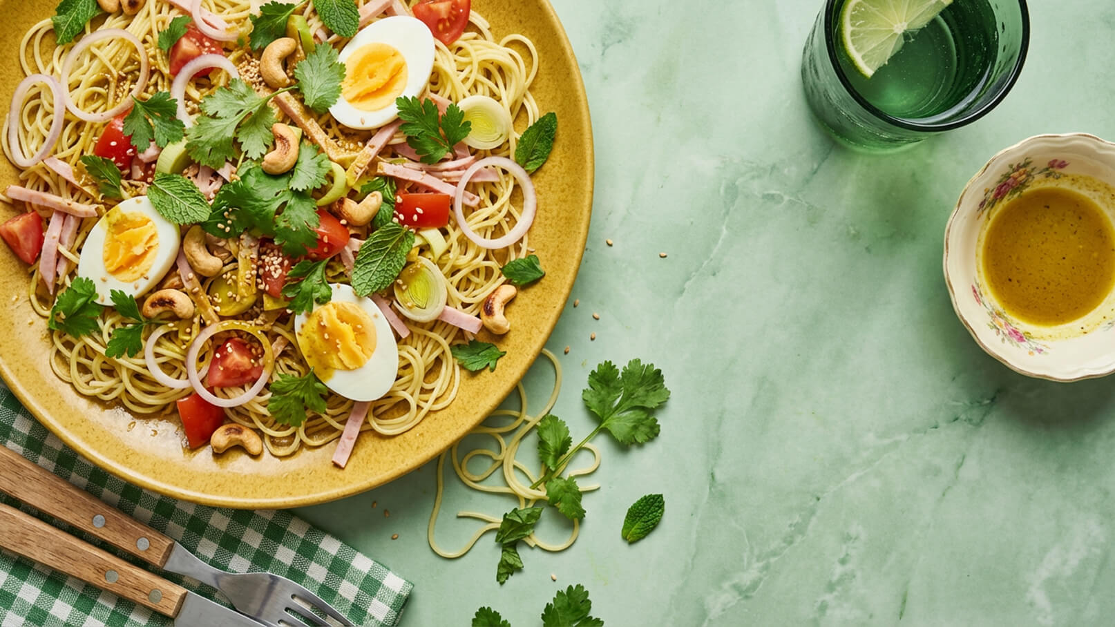Ein gelbes Pasta-Gericht mit Spaghetti, gekochten Eiern, Tomaten, Paprika, Lauch und Kräutern wie Koriander und Minze. Ergänzt mit Cashewkernen und Sesam. Daneben ein Glas mit Wasser und einer Limettenscheibe sowie eine kleine Schüssel mit einer gelben Soße.