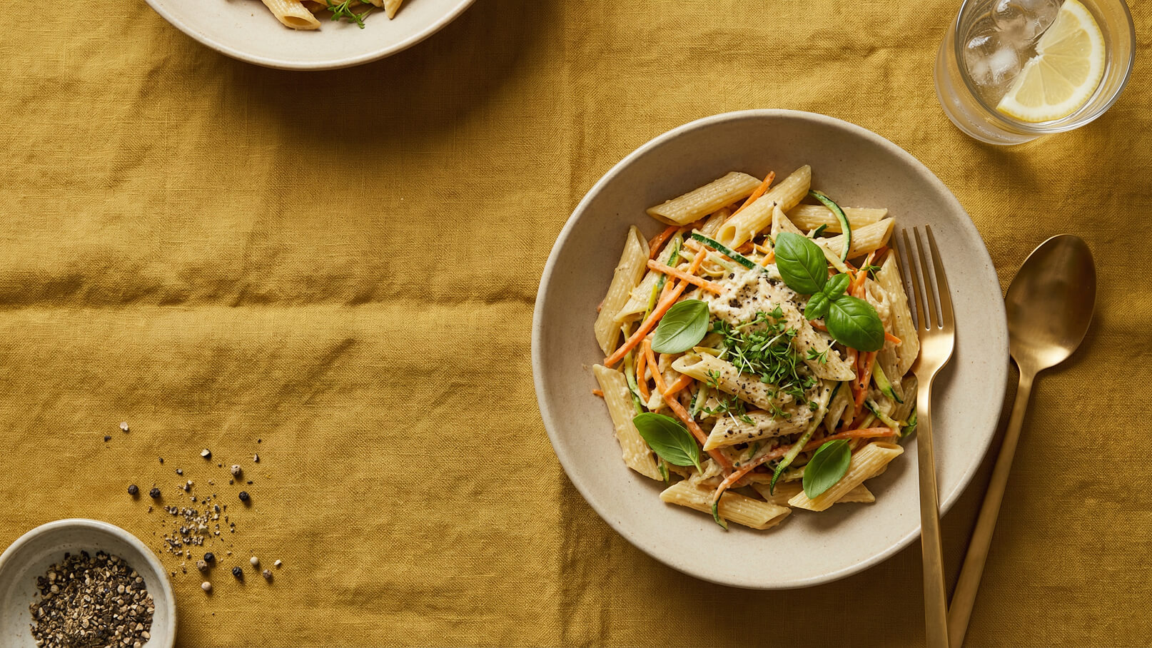 Ein Teller mit Penne-Nudeln, garniert mit frischen Kräutern und Gemüsestreifen, liegt auf einem gelben Tischtuch. Neben dem Teller befindet sich ein Essbesteck aus goldenem Metall. Ein Glas mit Wasser und einer Zitronenscheibe steht ebenfalls auf dem Tisch.