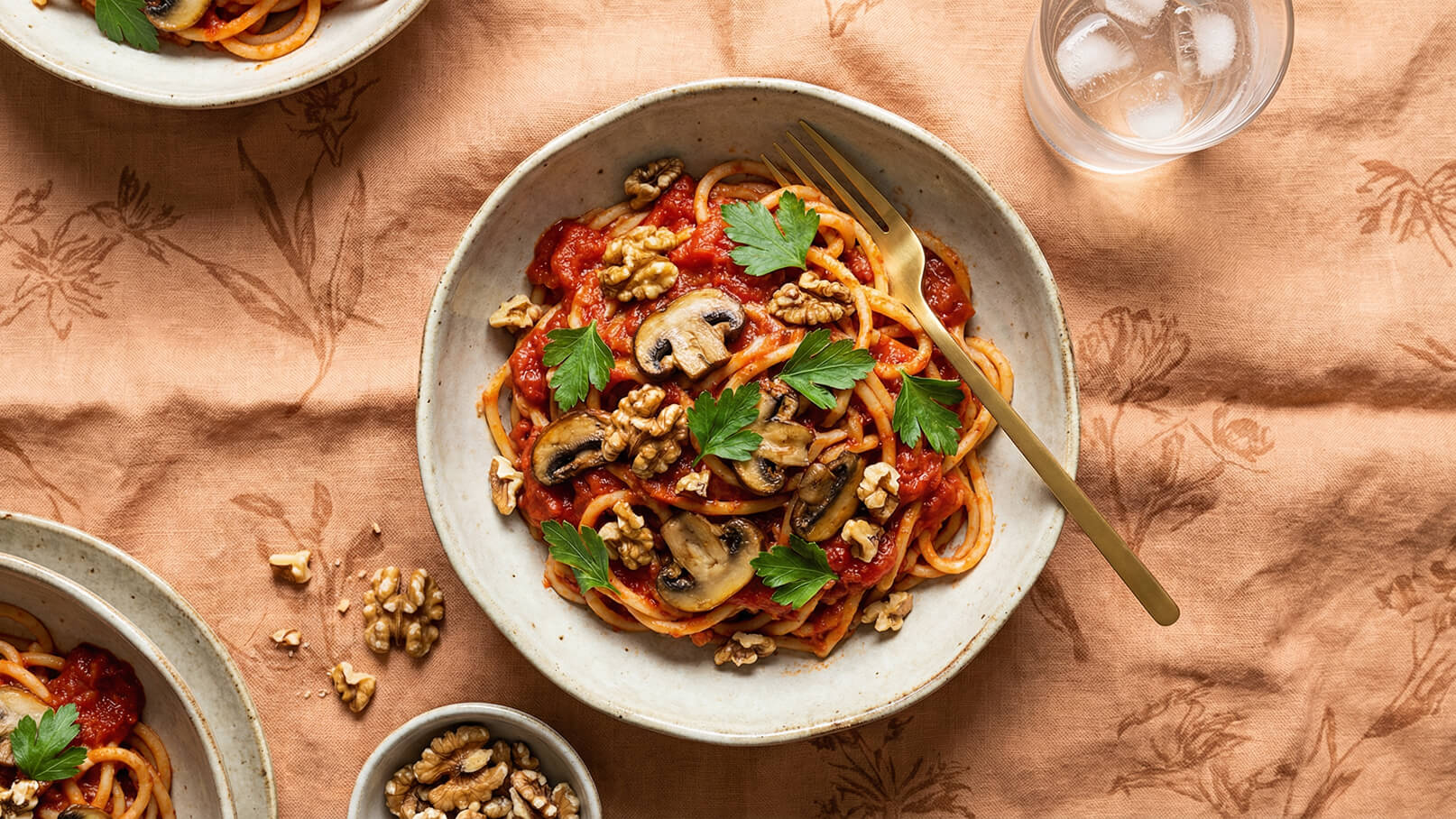 Ein Teller Spaghetti in Tomatensauce, garniert mit Champignons und Walnüssen, liegt auf einem neutralen Untergrund. Ein goldener Löffel steckt im Gericht. Neben dem Teller sind weitere Walnüsse und ein Glas mit Eiswasser sichtbar.