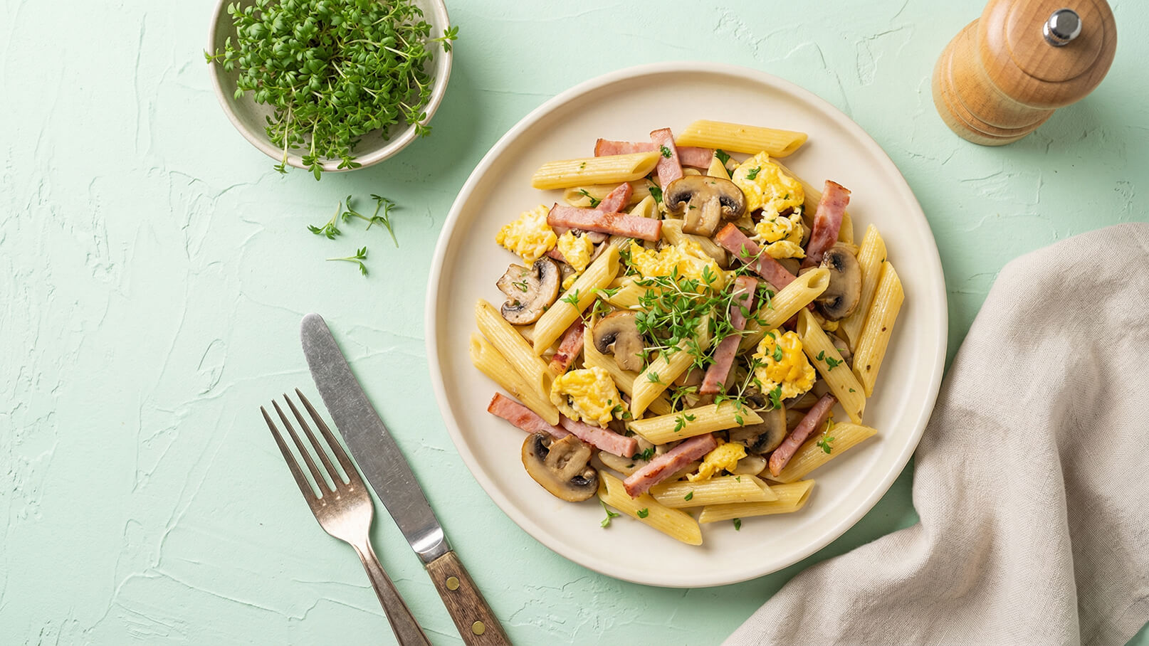 Ein Teller mit Penne-Pasta, garniert mit Rührei, Schinken und Champignons. Frische Kräuter liegen obenauf. Im Hintergrund sind eine Gabel und ein Messer sowie eine Schale mit grünen Kräutern zu sehen. Der Untergrund ist in sanften Minttönen gehalten.