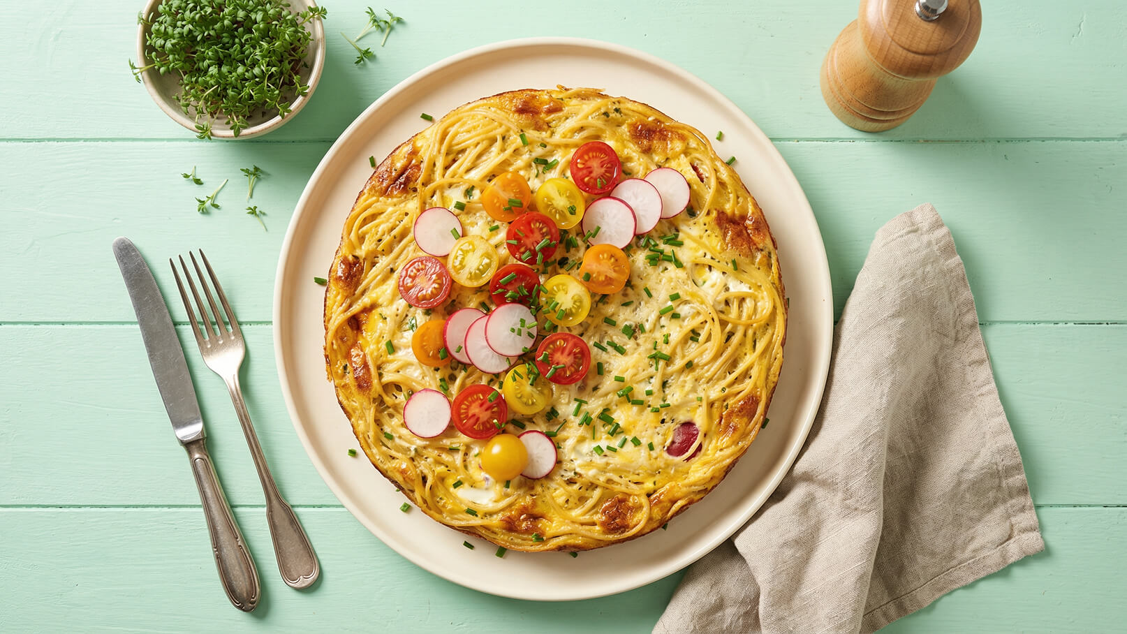 Ein runder Spaghetti-Auflauf liegt auf einem weißen Teller. Er ist mit bunten Kirschtomaten und Radieschen garniert, die auf der Oberfläche verteilt sind. Frischer Schnittlauch fügt Farbe hinzu. Eine Gabel und ein Messer liegen neben dem Teller, sowie ein Behälter mit frischen Kräutern im Hintergrund.