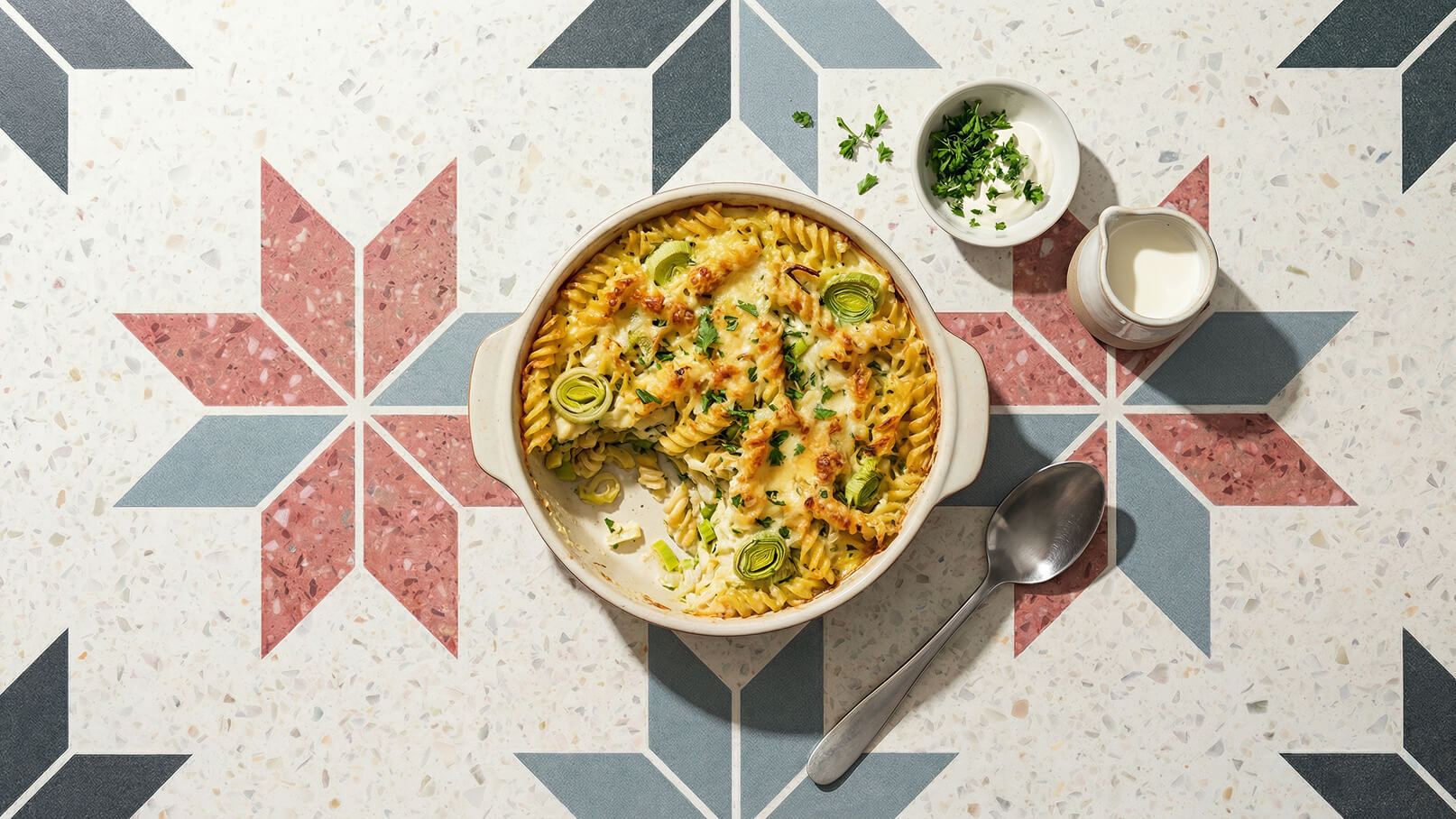 Ein metallischer Auflaufform mit gebackenem Pasta-Gericht, garniert mit Lauch und Petersilie, steht auf einem dekorativen Tisch mit geometrischem Muster. Neben der Form befinden sich eine kleine Schüssel mit gehackter Petersilie und eine Kanne mit einer weißen Soße.