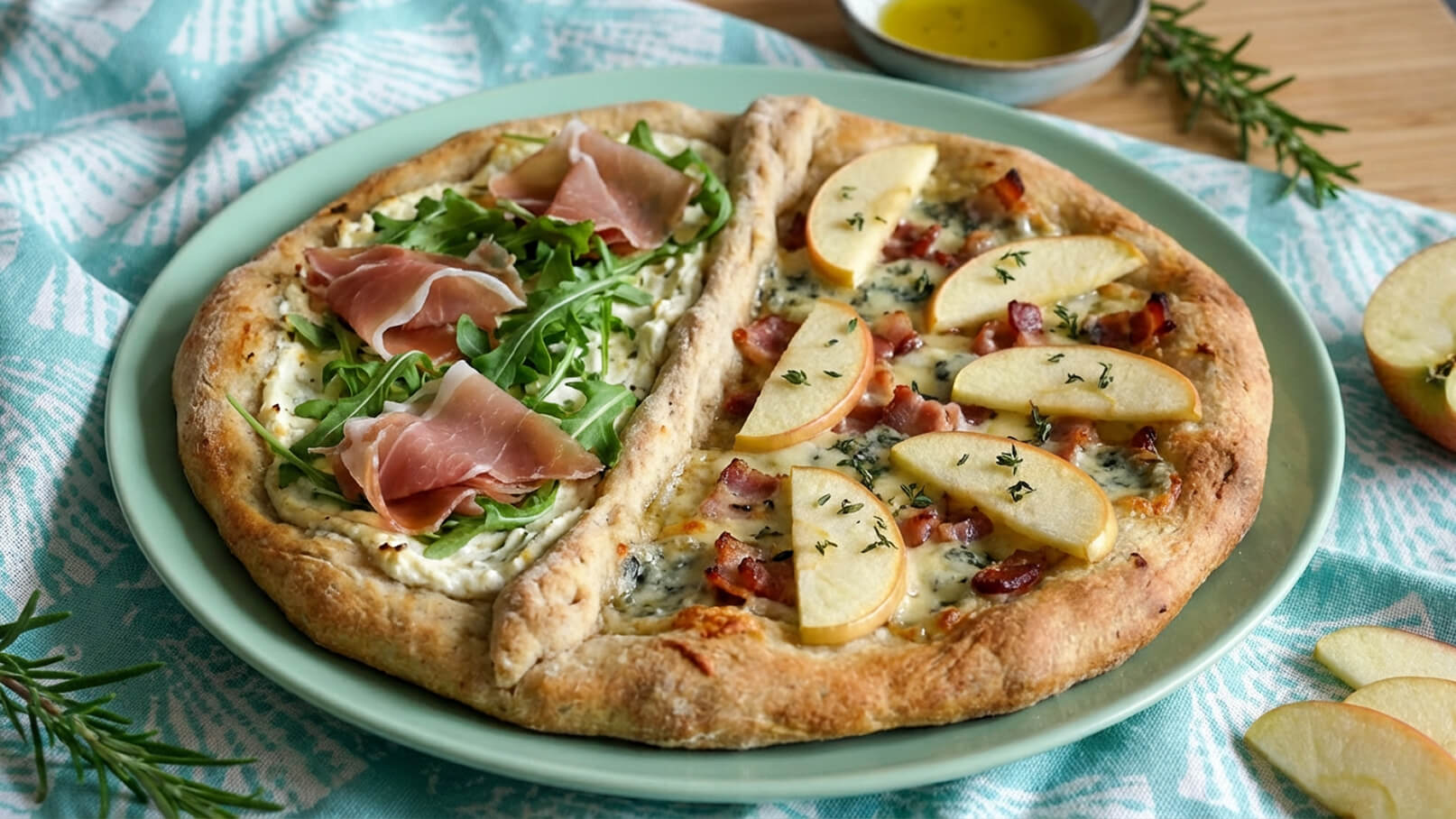 Auf einem grünen Teller liegen zwei Pizzastücke. Die linke Seite ist mit Rucola und Schinken belegt, während die rechte Seite Äpfel, Speck und Käse zeigt. Im Hintergrund ist ein kleiner Schälchen mit Olivenöl und frischem Rosmarin zu sehen.