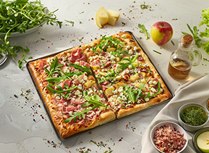 Eine rechteckige Pizza mit vier verschiedenen Belägen, darunter Schinken, Ananas und Rucola. Sie liegt auf einem Backblech und ist umgeben von frischen Zutaten wie einem Apfel, einer Ölpumpe und diversen Gewürzen.