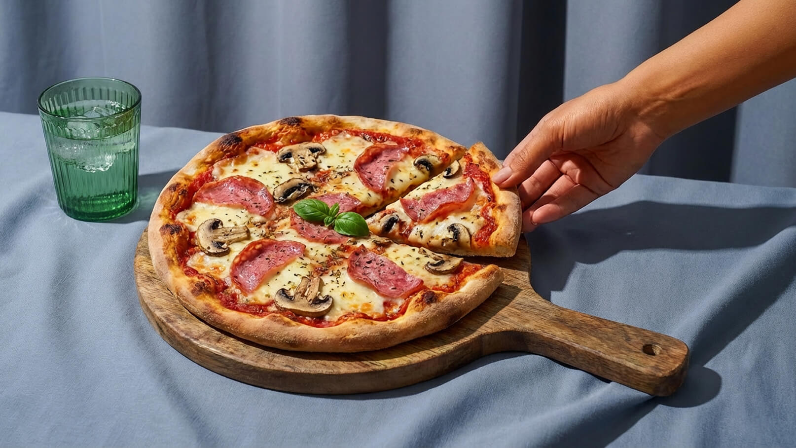 Ein runder Pizzateller aus Holz mit einer frisch gebackenen Pizza, belegt mit Käse, Salami, Schinken und Champignons. Eine Hand greift nach einem Stück. Im Hintergrund steht ein Glas mit Wasser. Der Tisch hat eine graue Tischdecke.