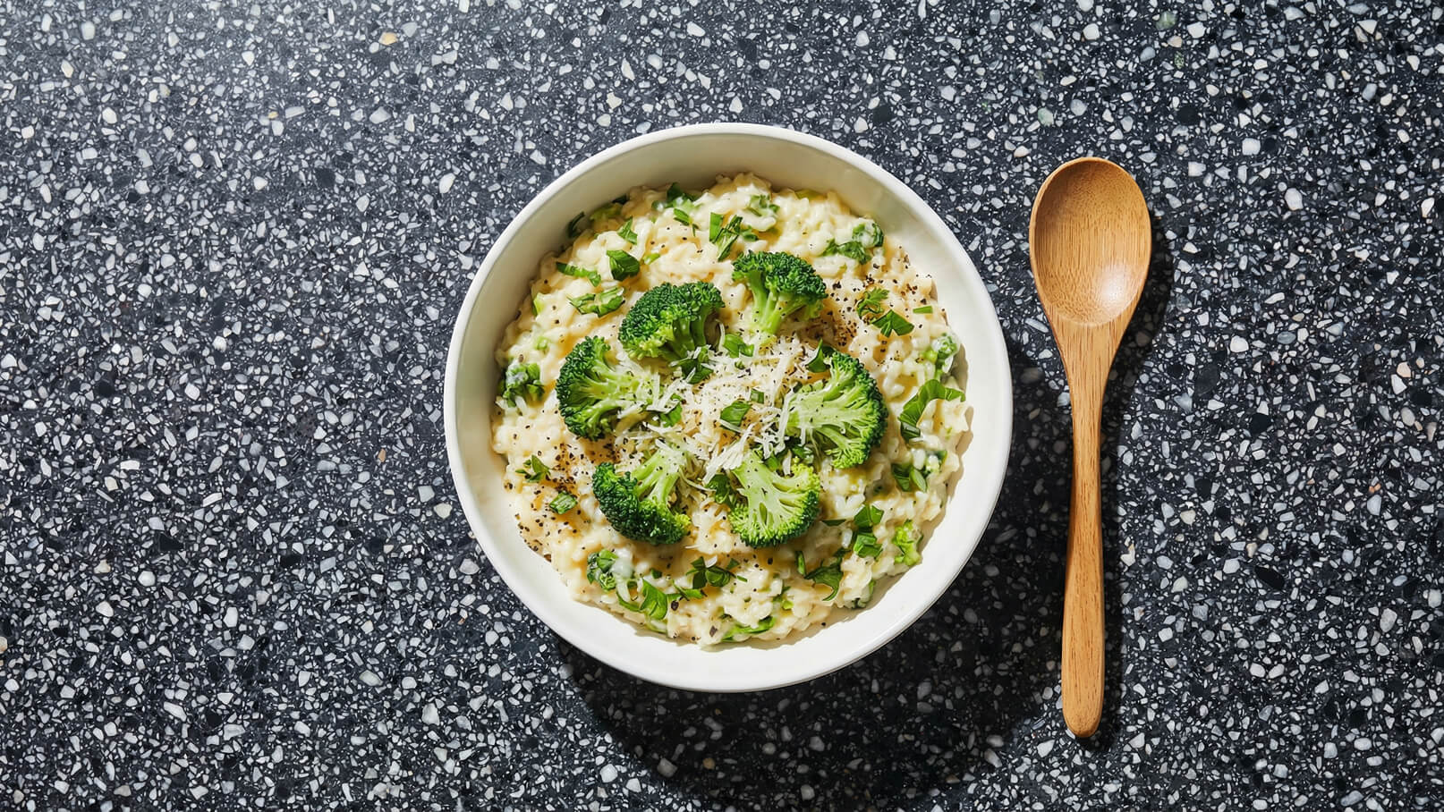 Ein Teller Risotto mit grünen Erbsen und Brokkoli, garniert mit frisch geriebenem Käse und Kräutern. Der Teller ist auf einer strukturierten, dunklen Oberfläche platziert, neben dem Teller liegt ein Holzlöffel.