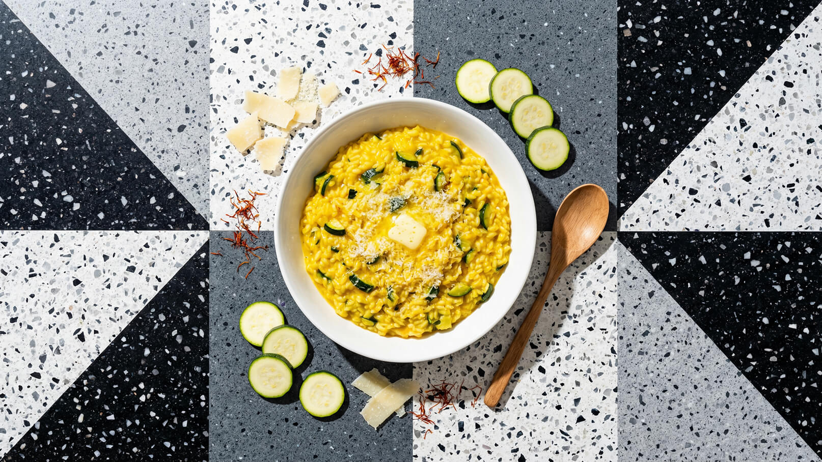 Ein weißer Teller mit cremigem Risotto, garniert mit Zucchinistücken und einem Stück Butter in der Mitte. Neben dem Teller liegen einige Käsestreifen, Zucchinischeiben und ein Holzlöffel. Der Hintergrund zeigt ein geometrisches Muster aus schwarzen und grauen Fliesen.
