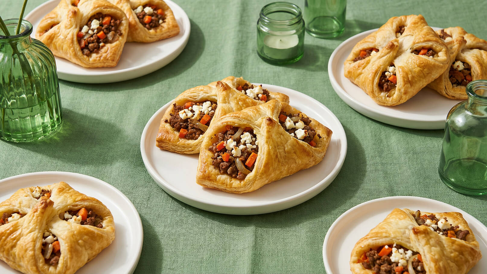 Auf dem Bild sind mehrere gefüllte Blätterteigtaschen auf weißen Tellern zu sehen. Die Füllung besteht aus Hackfleisch, Karotten und Käse. Der Hintergrund ist ein grüner Tisch, auf dem auch einige Gläser stehen.