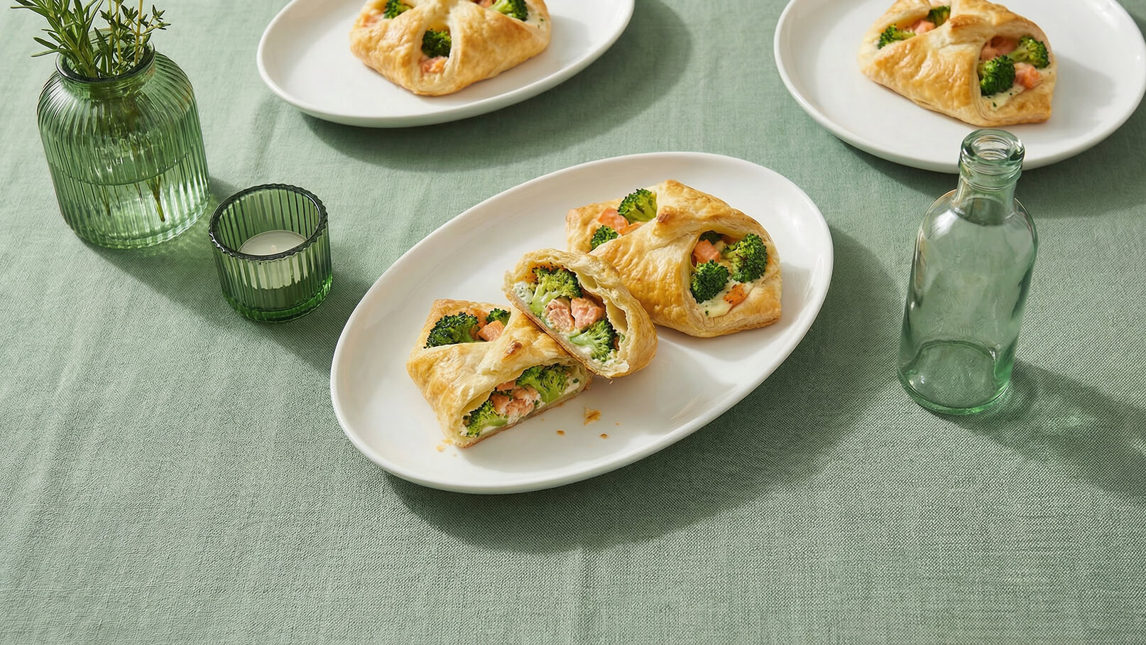 Auf einem weißen Teller liegen gefüllte Teigtaschen mit Brokkoli und Lachs. Eine der Teigtaschen ist aufgeschnitten, sodass die Füllung sichtbar ist. Der Hintergrund ist ein weißes Tuch auf einem grünen Tisch.