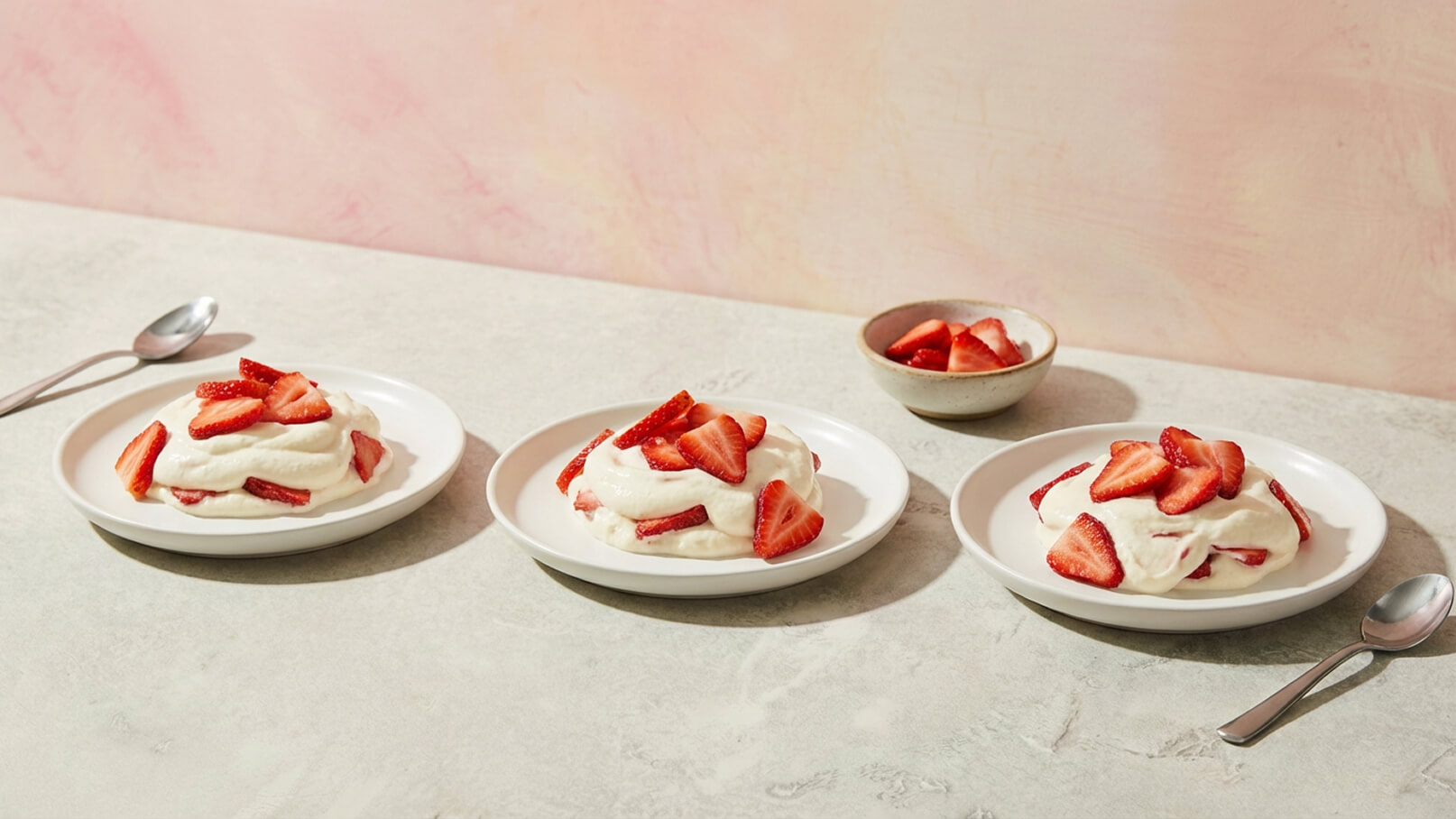 Drei Teller mit einer Sahne-Nachspeise sind auf einem Tisch angeordnet. Jeder Teller enthält eine Portion mit geschlagener Sahne und frischen Erdbeeren. Im Hintergrund befindet sich eine kleine Schüssel mit zusätzlich geschnittenen Erdbeeren. Der Tisch ist hell und schlicht.