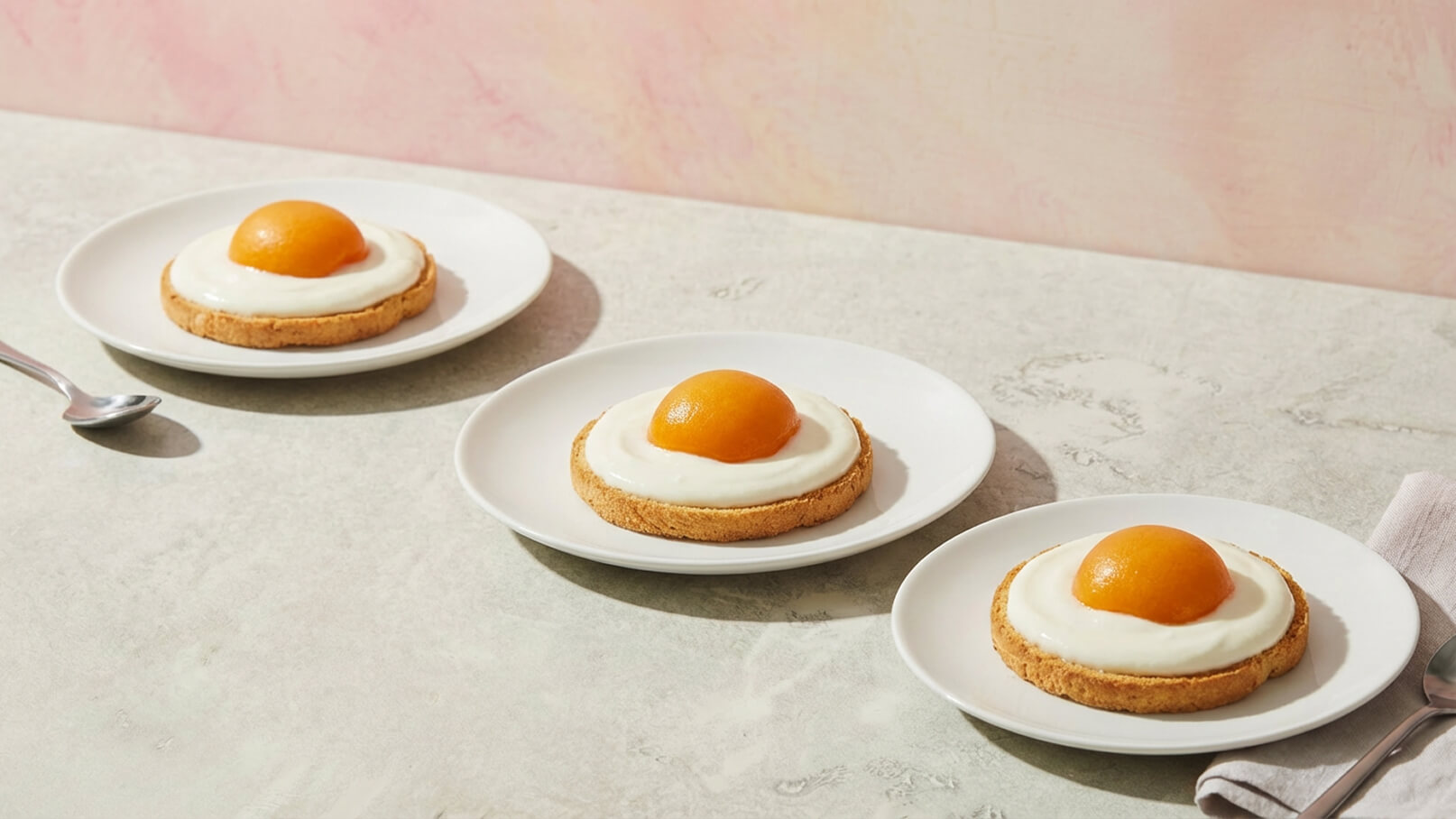 Drei Teller mit jeweils einem Keksboden, der mit einer dicken Schicht Joghurt oder Sahne bedeckt ist. In der Mitte jedes Tellers befindet sich eine orangefarbene Frucht, die einem Eigelb ähnelt. Der Hintergrund ist hellrosa.