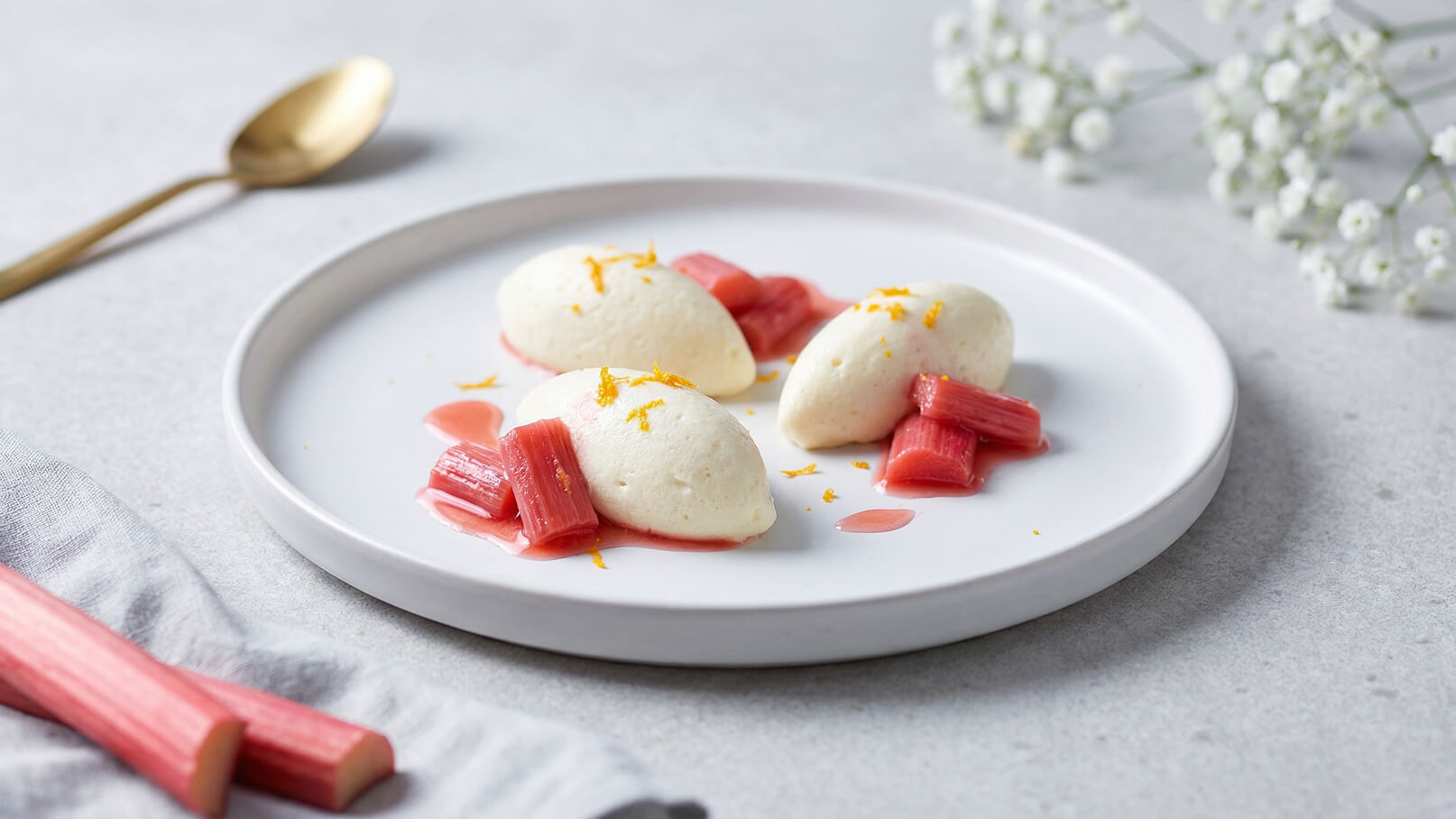 Ein eleganter Teller mit drei Portionen einer cremeartigen Nachspeise, begleitet von leicht rotem Rhabarber und einer fruchtigen Sauce. Die Nachspeise ist mit feinen Orangenstückchen garniert, die dem Gericht eine frische Note verleihen.