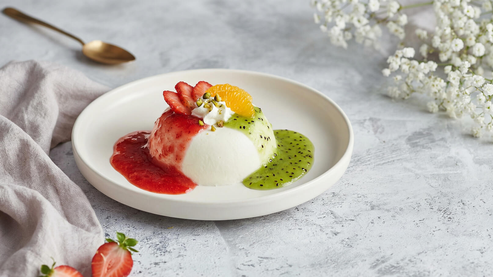 Ein Teller mit Panna Cotta, verziert mit Erdbeeren, einer Scheibe Orange und Pistazien. Die Panna Cotta wird von rotem Erdbeersirup und grünem Kiwisauce umgeben. Im Hintergrund sind ein Löffel und einige Blüten sichtbar.
